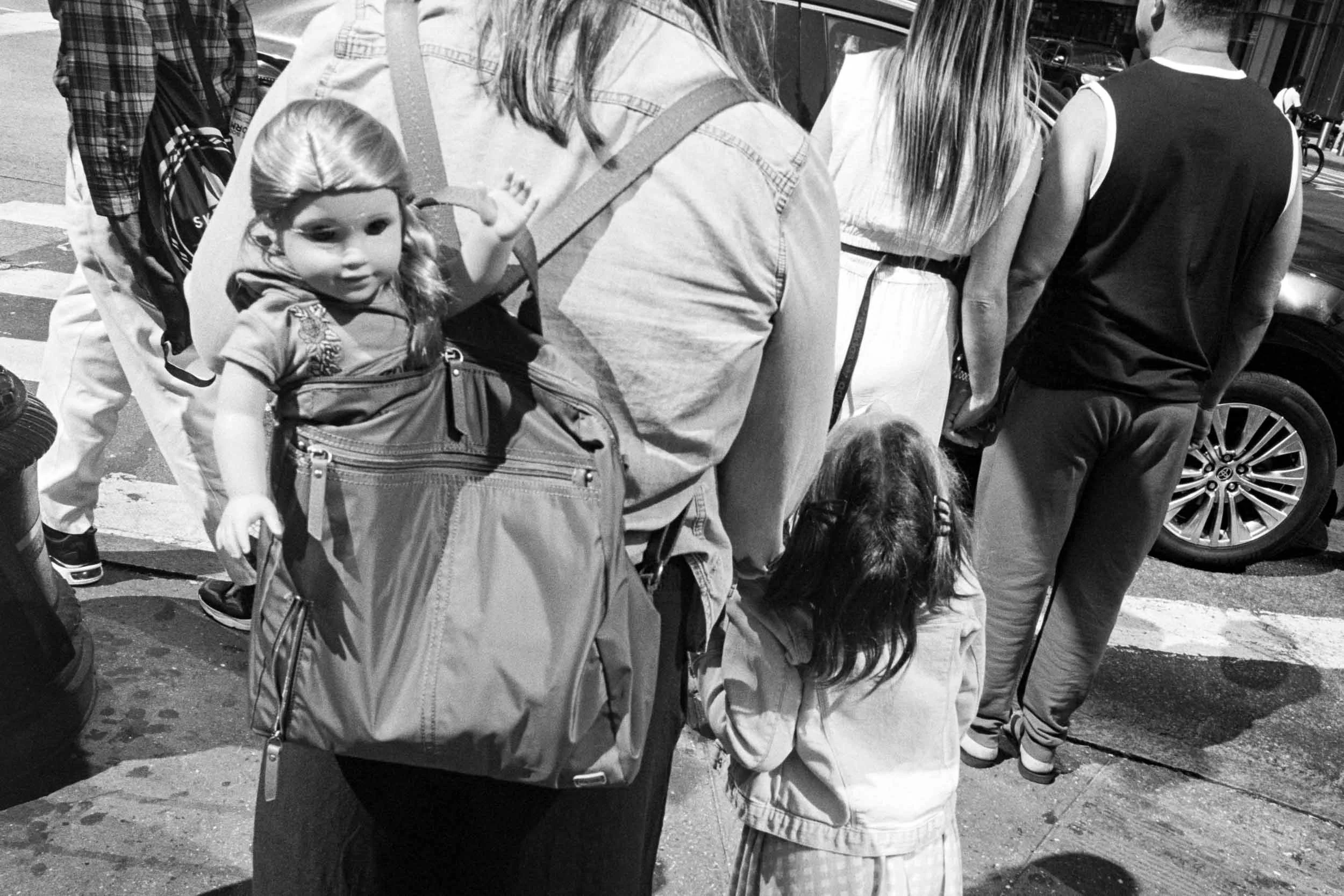 A picture taken at Bryant Park in New York f a woman and child waiting to cross the road. The woman has a backpack on with a doll sticking out.