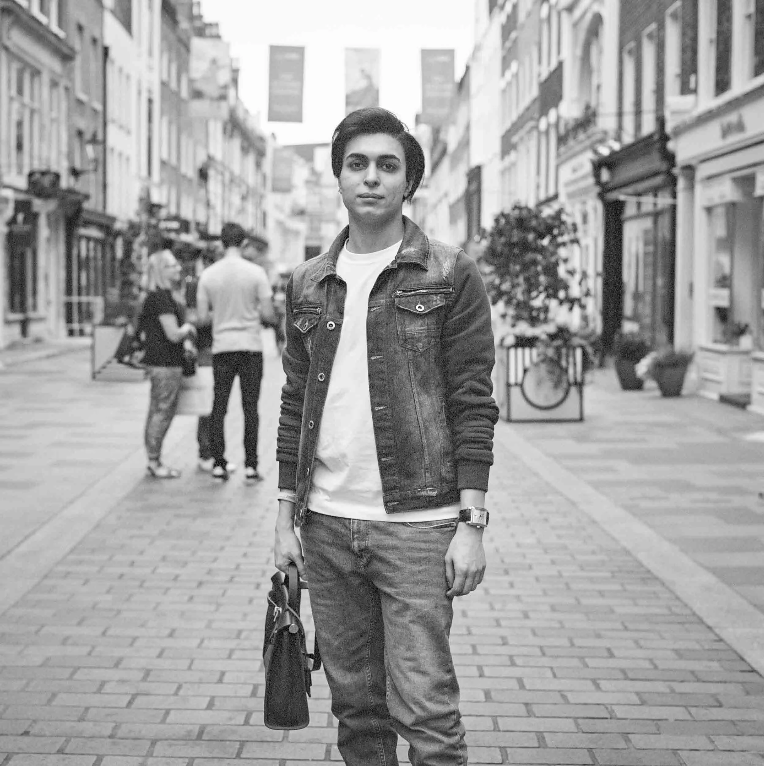 A person with short dark hair wearing a leather jacket and jeans walking down a city street with shops on either side, holding a bottle in one hand.