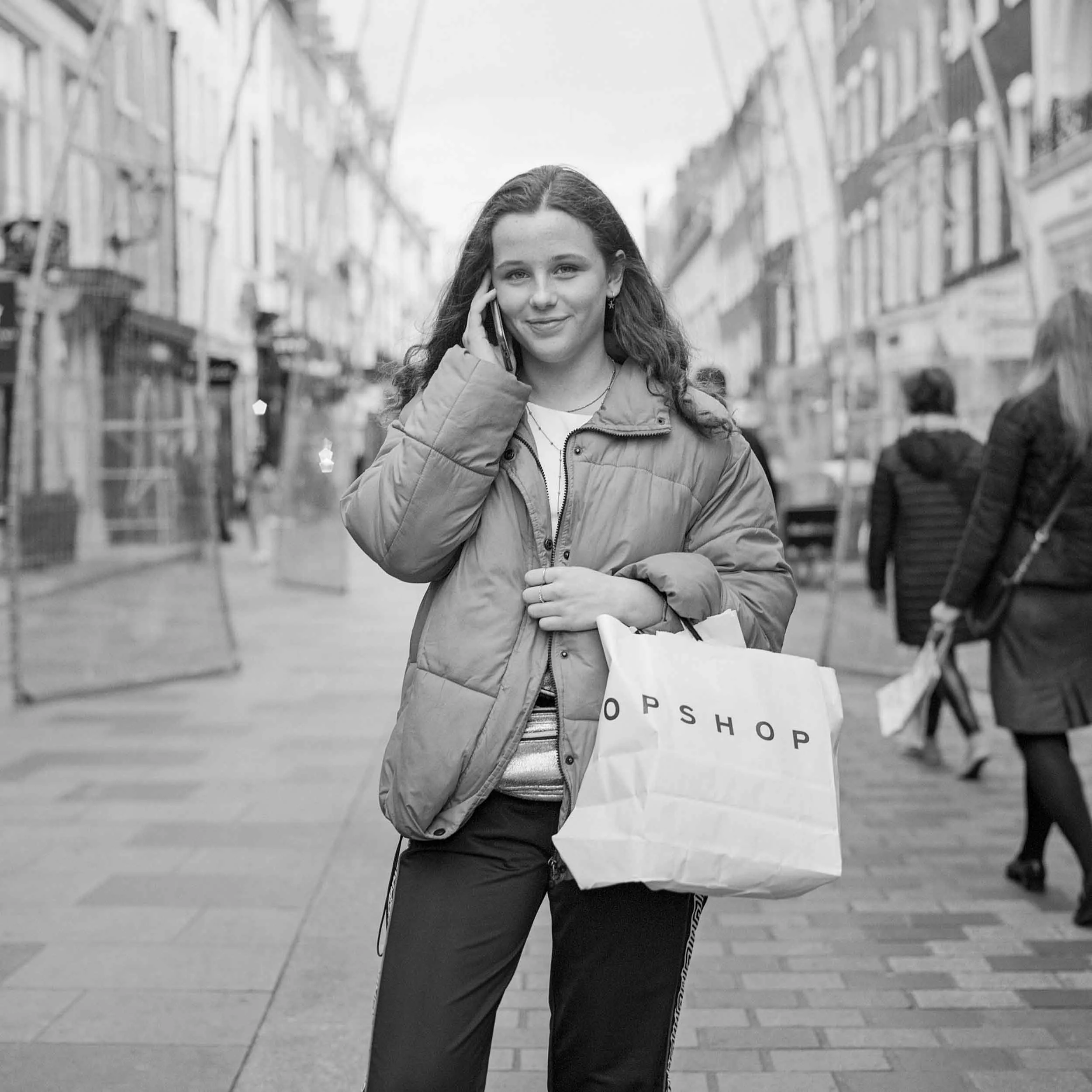 A young woman smiling and talking on her cellphone while walking on a city street, carrying a shopping bag, wearing a jacket and casual pants, with buildings and other pedestrians in the background.