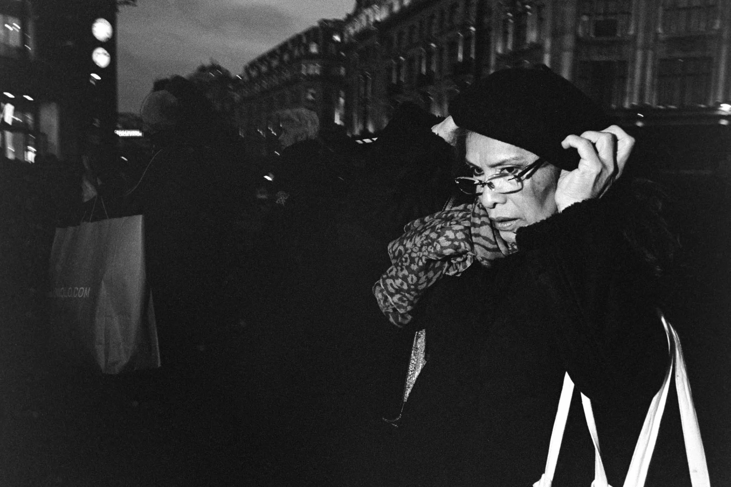 A person with glasses putting on a winter hat in an outdoor urban area at night, with buildings and a clock tower in the background.