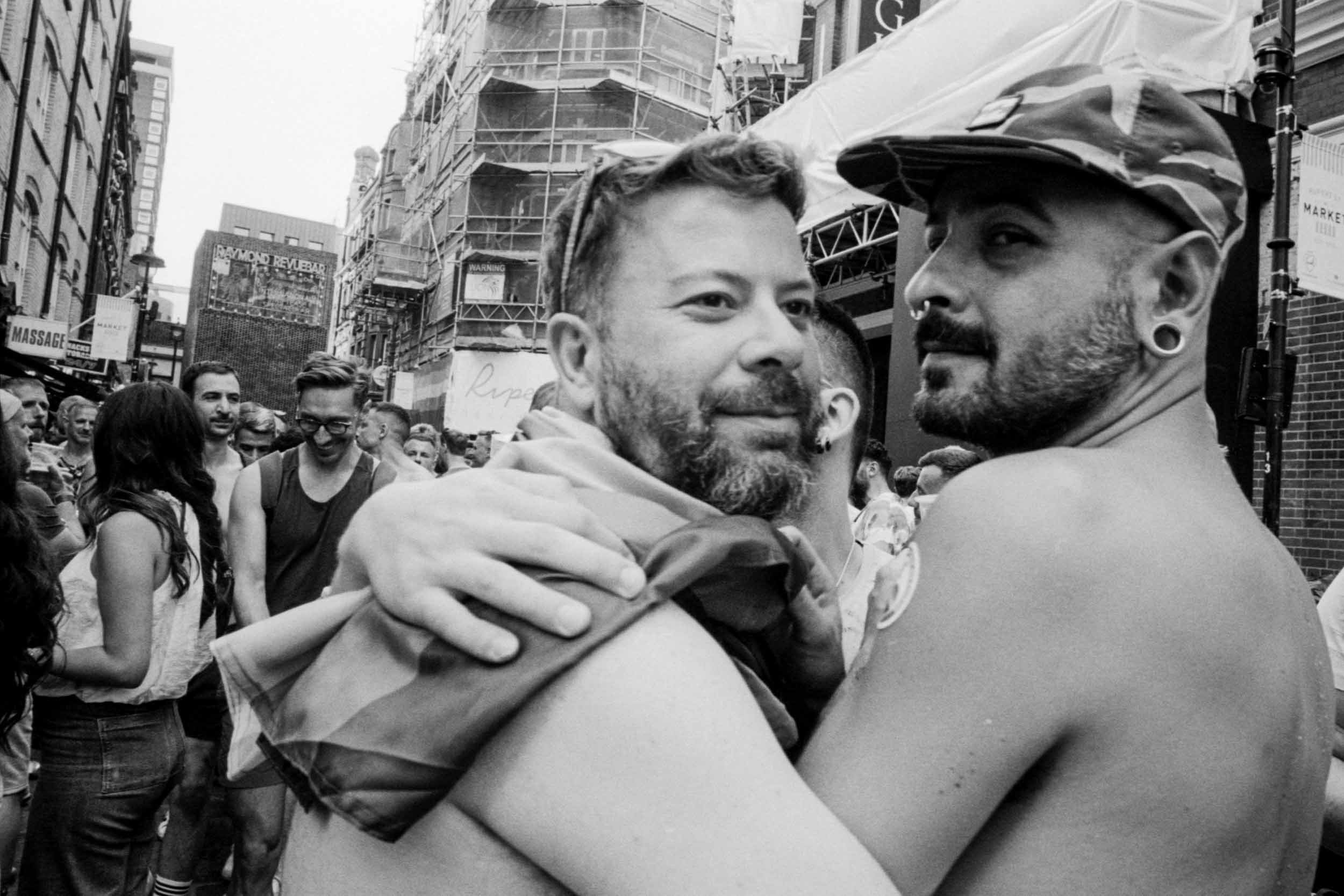 Two men hugging each other in a busy city street with buildings, signs, and people in the background.