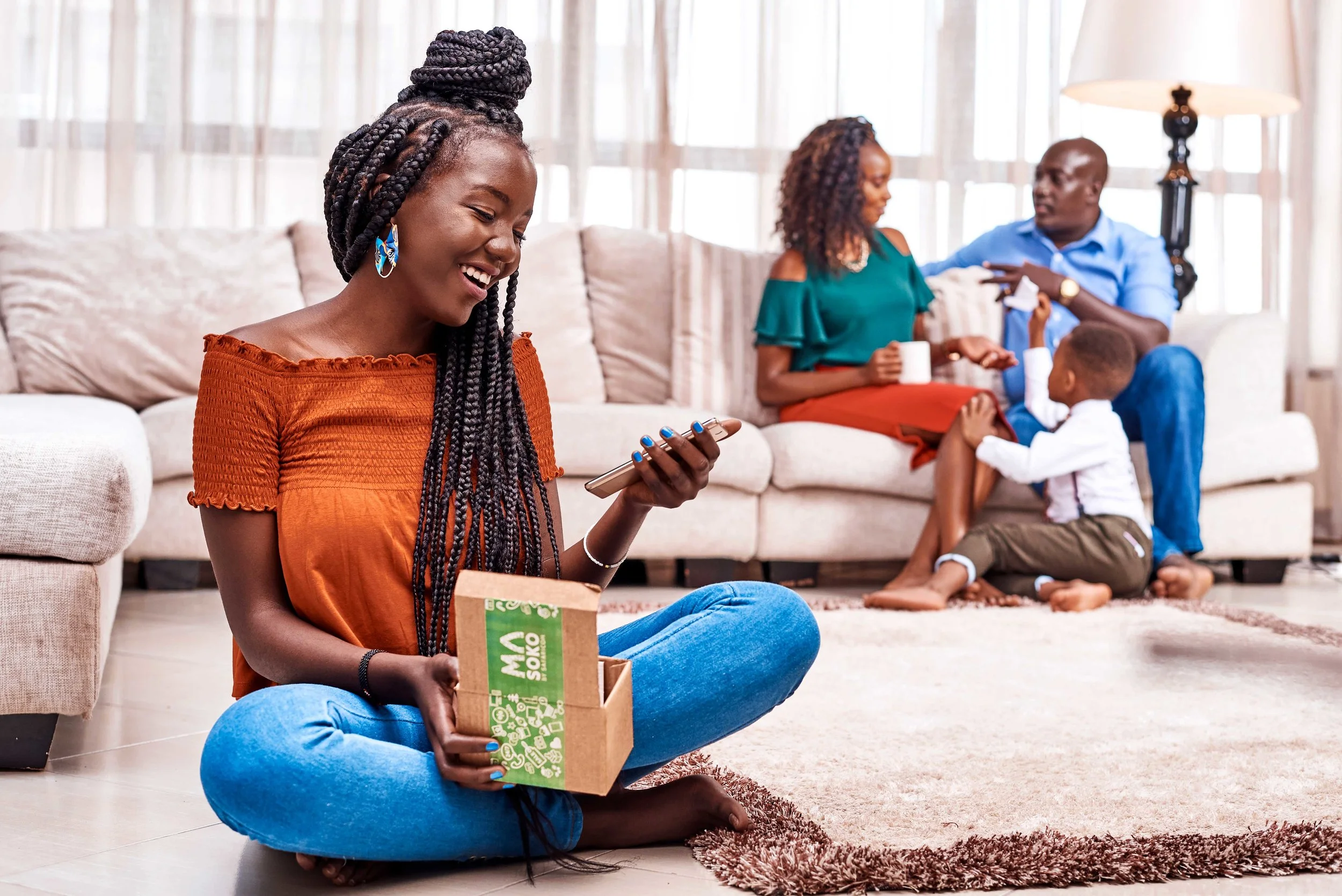 A young woman sitting on the floor with a smartphone and an open box, smiling. In the background, a family sits on a couch; a man and woman talk while a child plays.