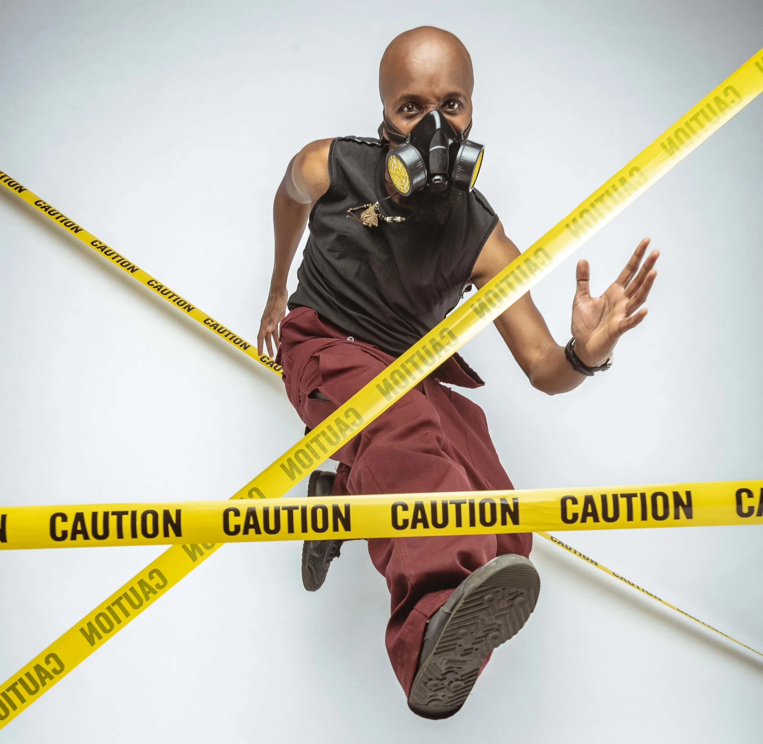 Person in a gas mask jumping through yellow caution tape, wearing a black tank top and red pants, against a white background.
