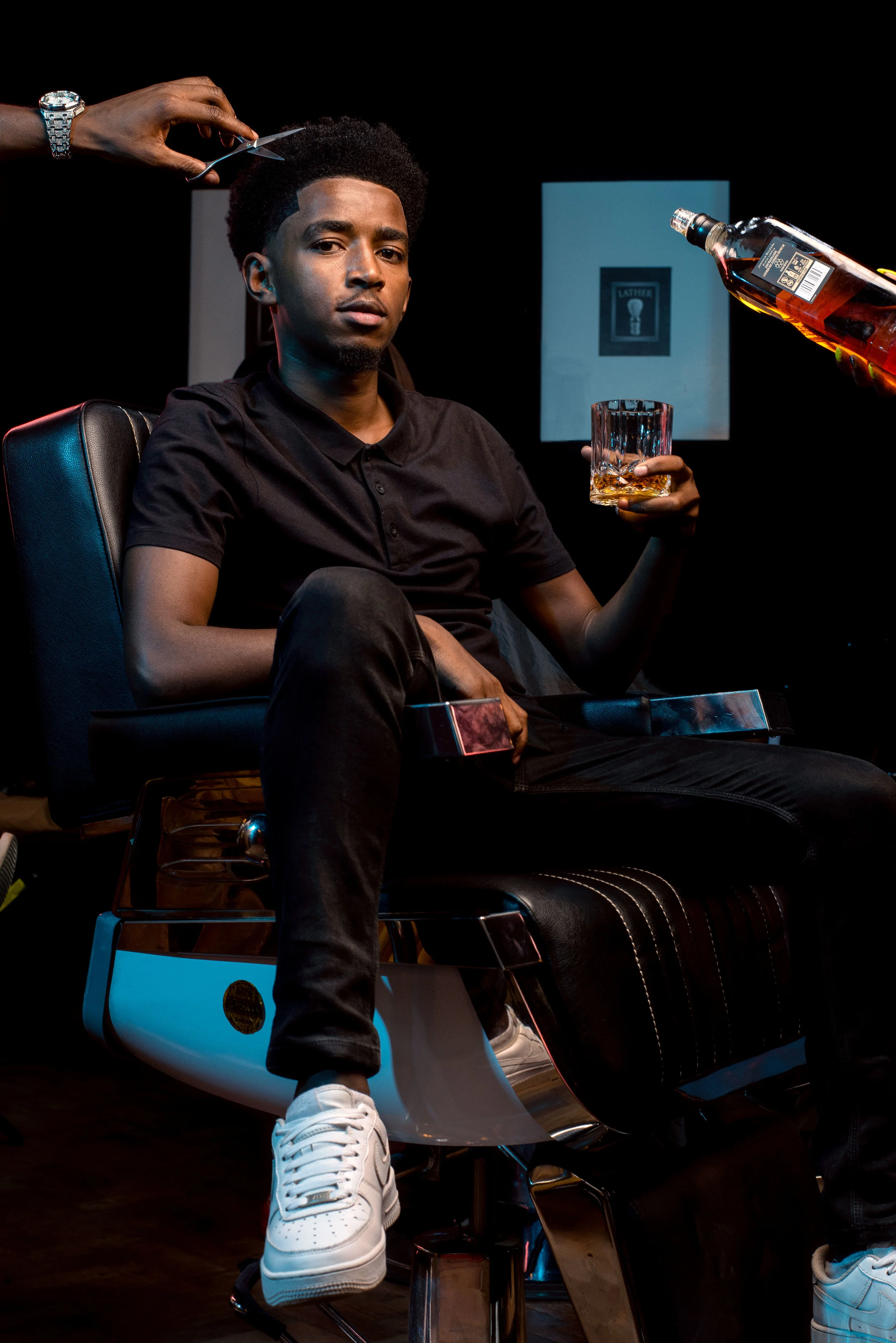 Man sitting in a barber chair getting a haircut, holding a glass of whiskey, with another hand pouring whiskey.