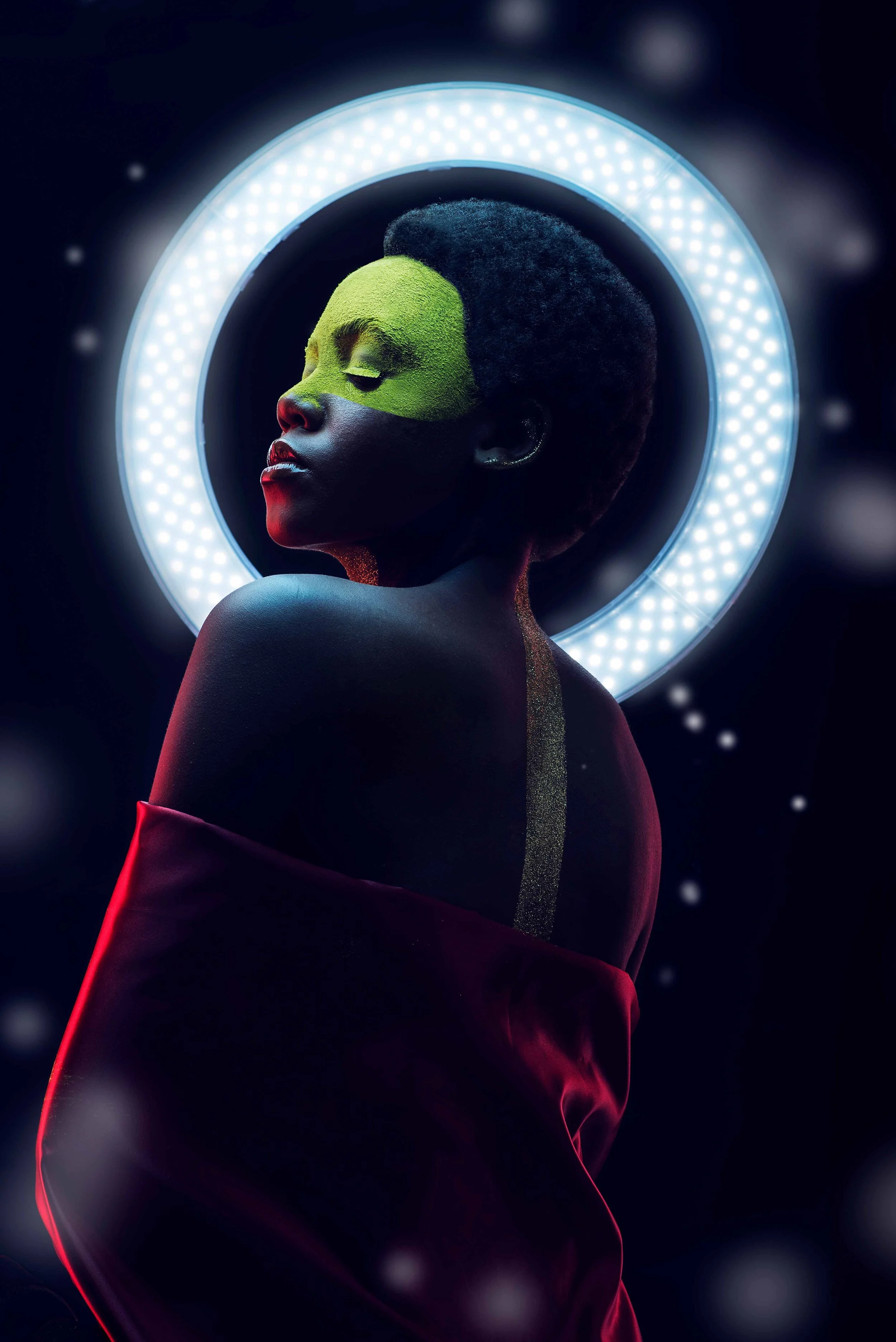 Artistic portrait of a person with green makeup, illuminated by a circular LED light, with a dark background.