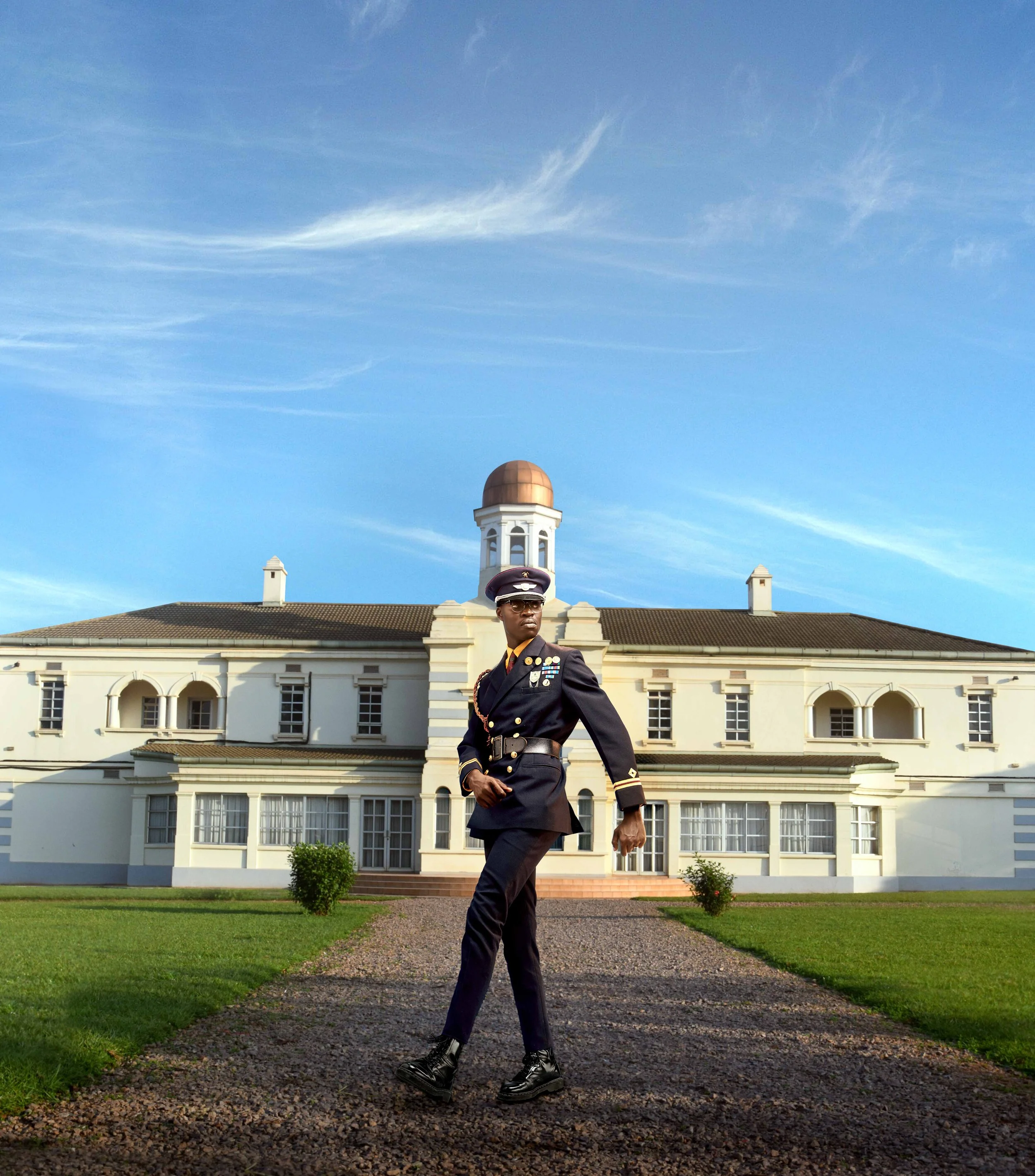 Person in a formal military uniform walking on a path in front of a historic building with a dome and a clear blue sky.