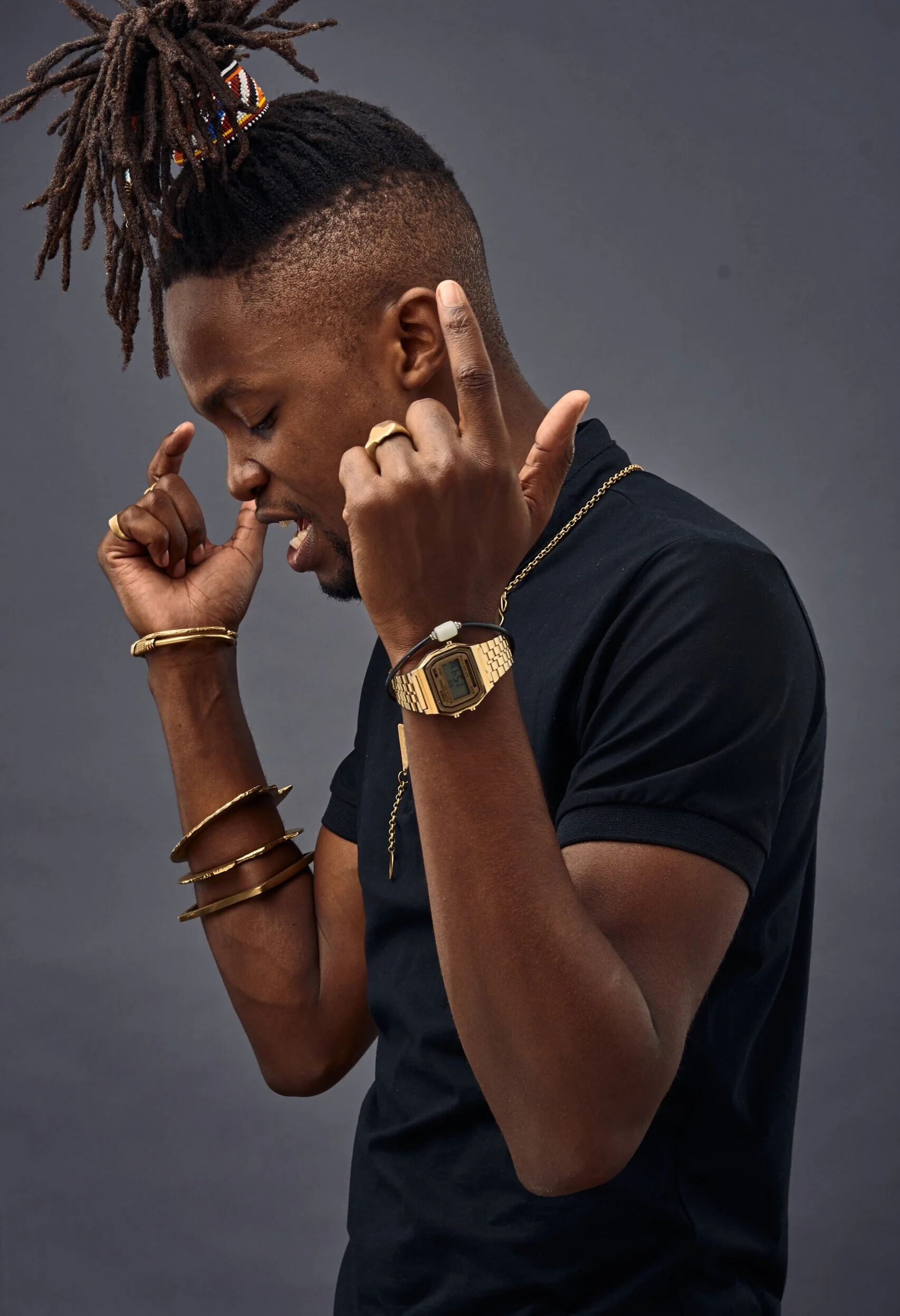 Person with dreadlocks, wearing gold jewelry and a black shirt, holding fingers near face against gray background.