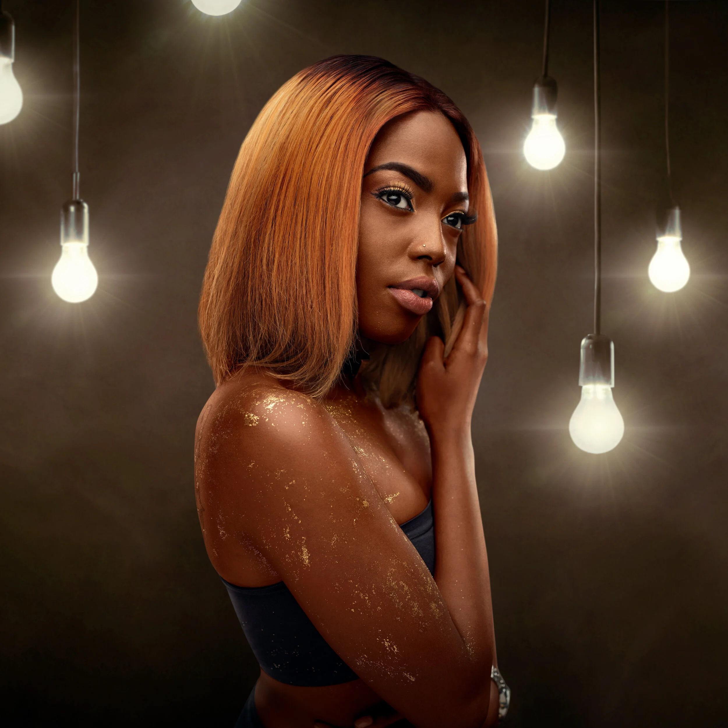 Portrait of a woman with golden skin glitter and shoulder-length hair, surrounded by glowing light bulbs in a dark setting.