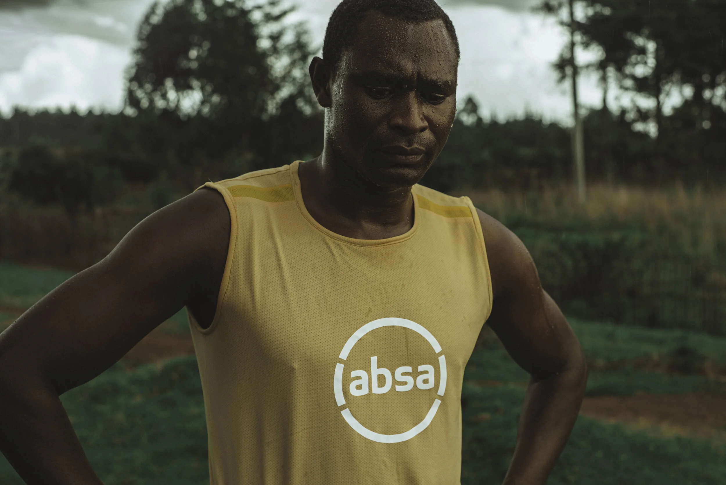 David Rudisha wearing a yellow tank top with "absa" logo, standing outdoors, looking down, with trees in the background.