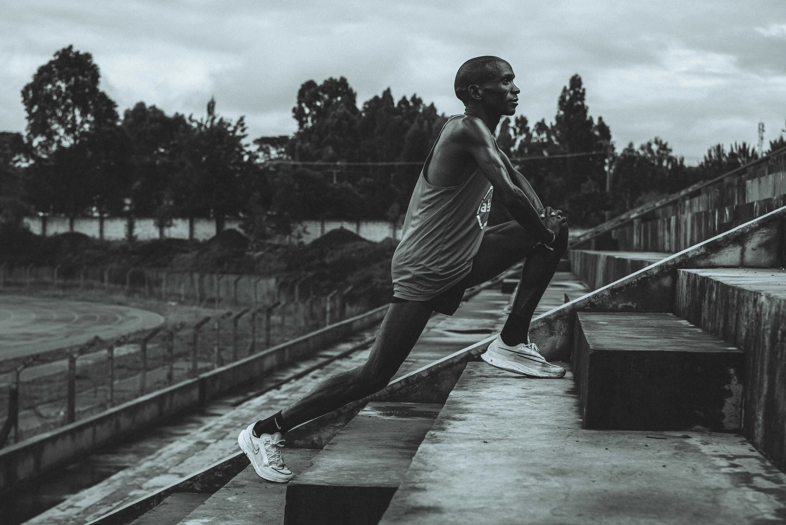 Eliud Kipochoge stretching on an outdoor stadium staircase wearing athletic gear and sneakers, with a track and trees in the background.