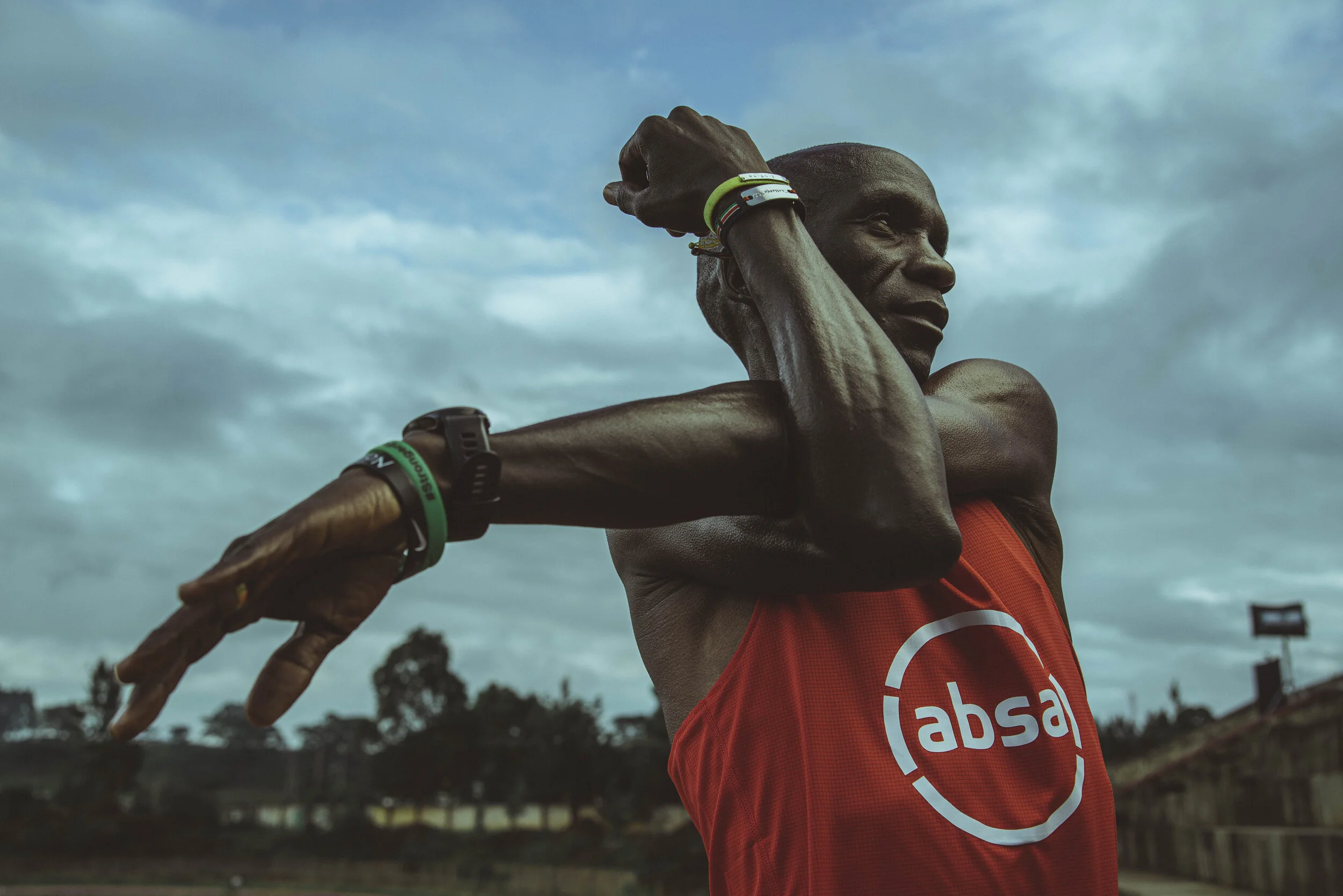 Athlete stretching outdoors in a red shirt with "absa" logo, wearing wristbands and a watch.