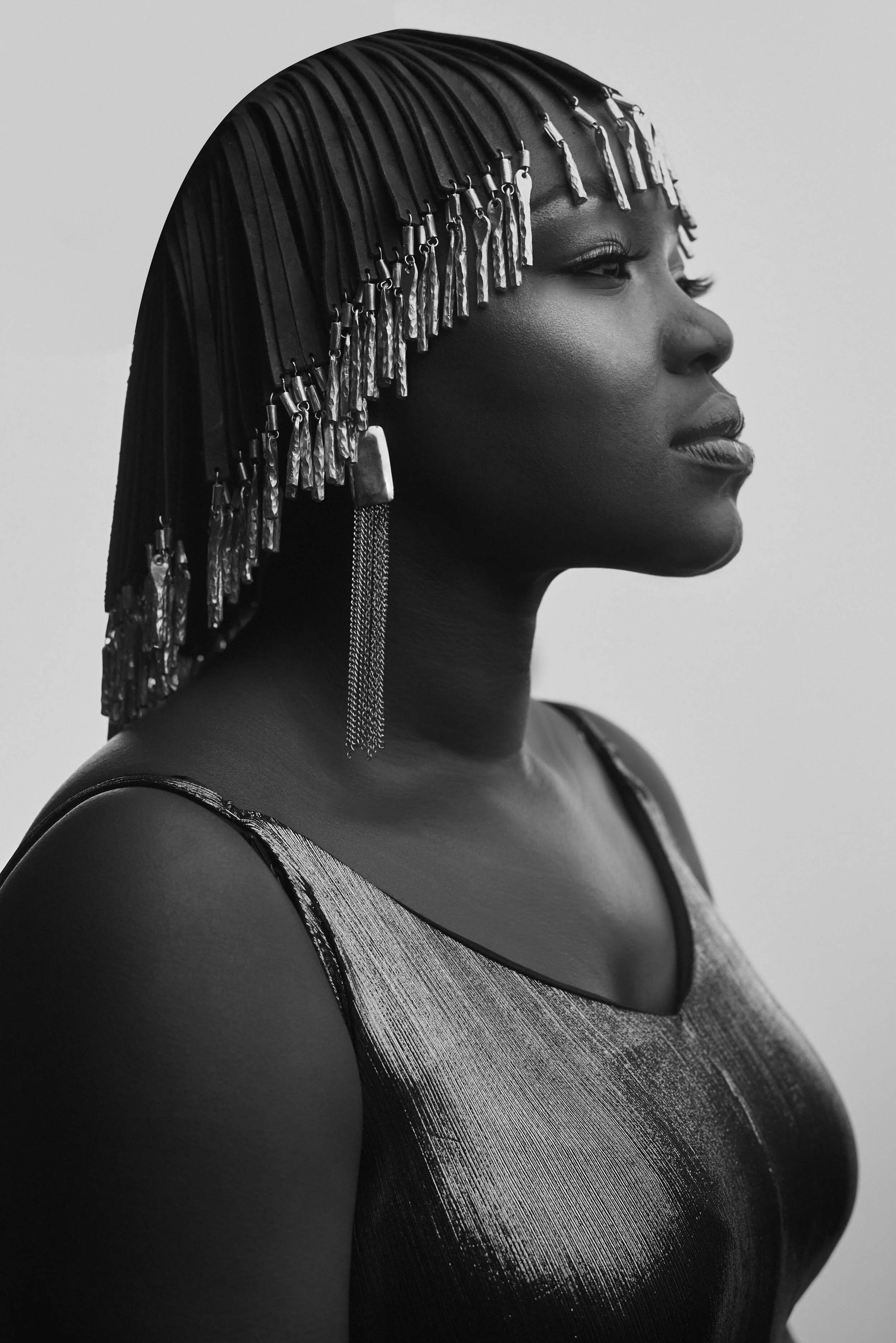 Black and white portrait of a person wearing a headpiece with fringe and statement earrings, dressed in a shiny textured top, side profile view.