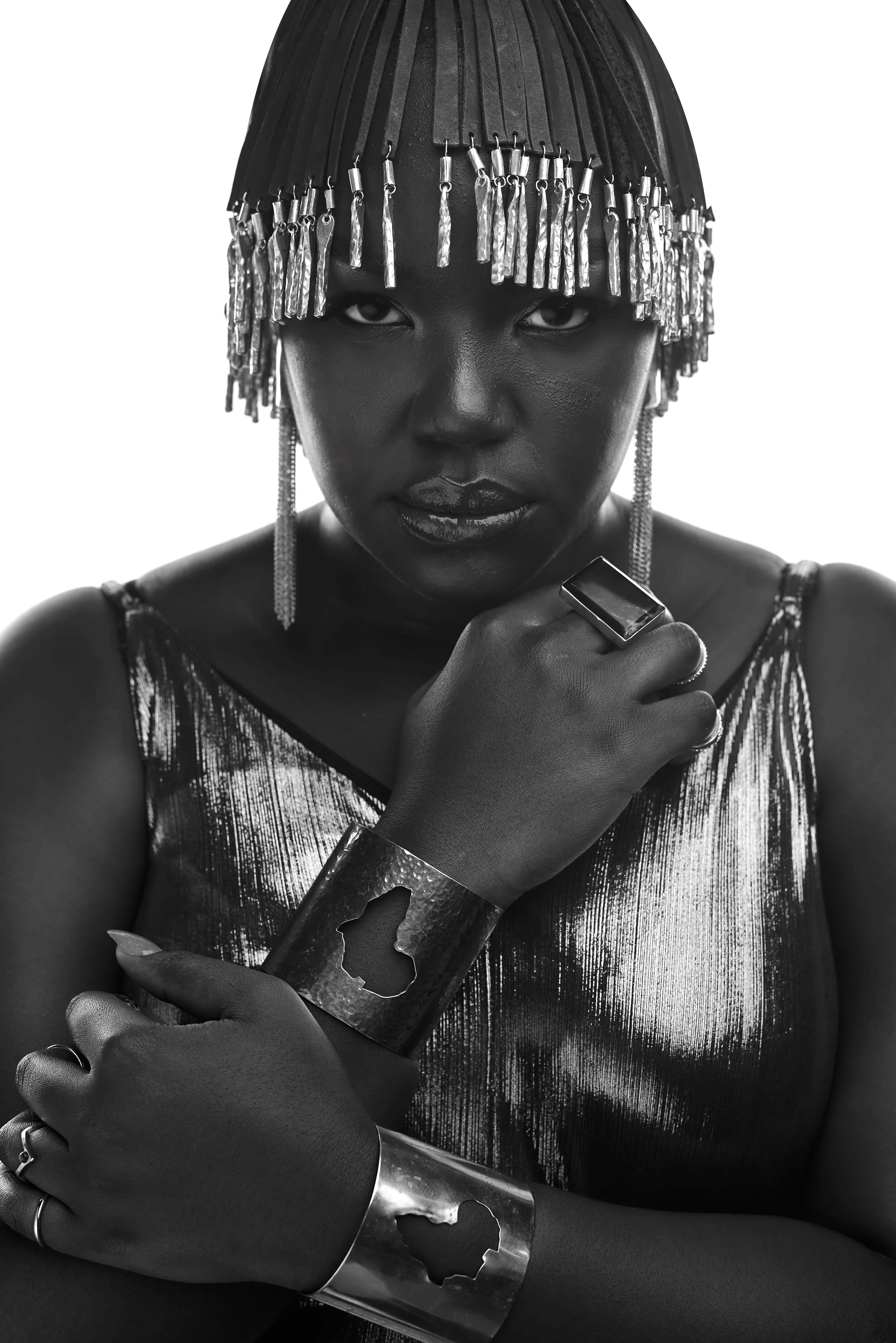 Black and white portrait of a person wearing a fringed headpiece, metallic jewelry, and a shiny top.