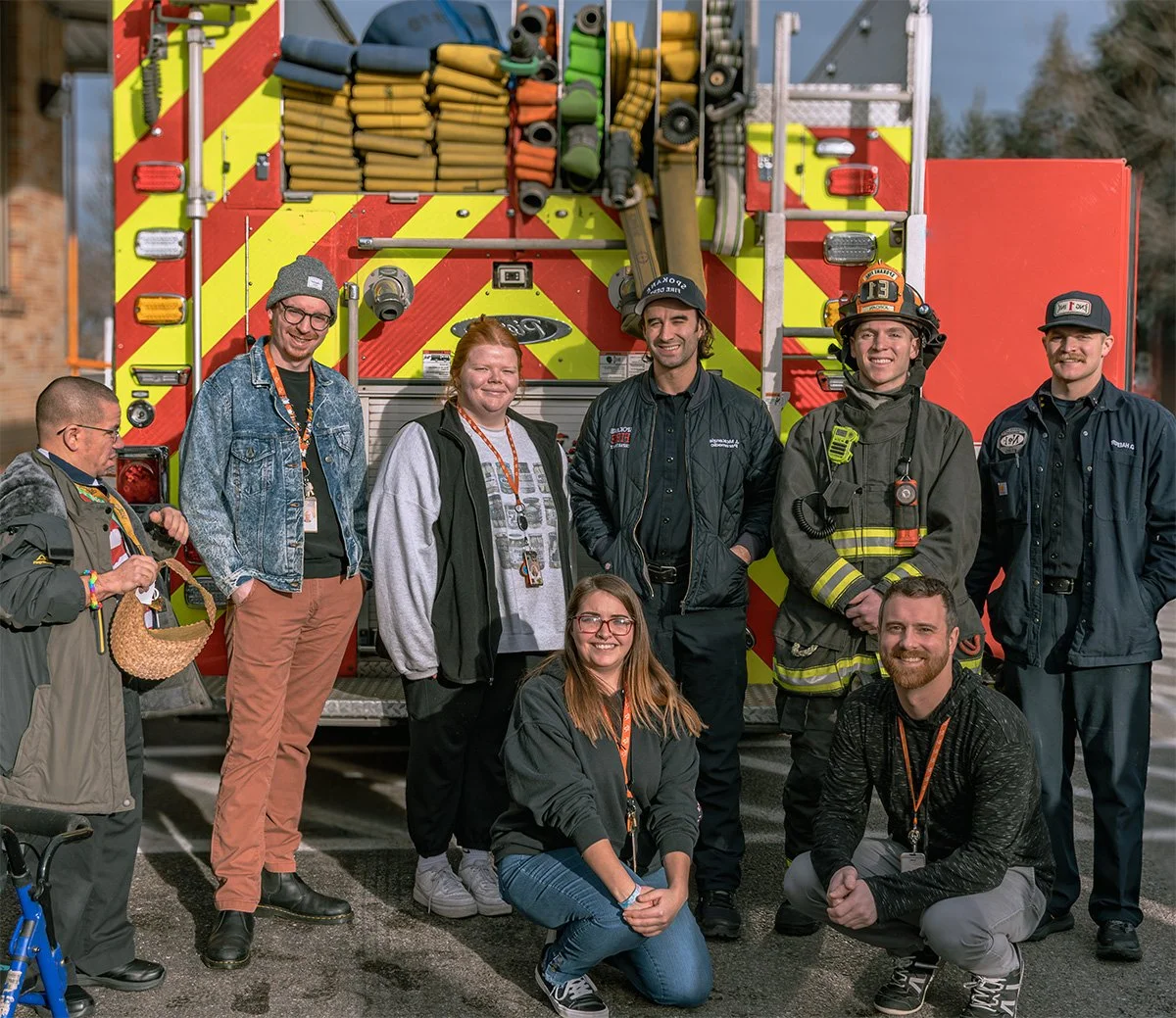 The Arc of Spokane staff smile alongside three District 13 firefighters in front of a fire engine. The group is arranged in a friendly semi-circle on a sunny day at the Community Center. On-screen text identifies The Arc of Spokane staff members.