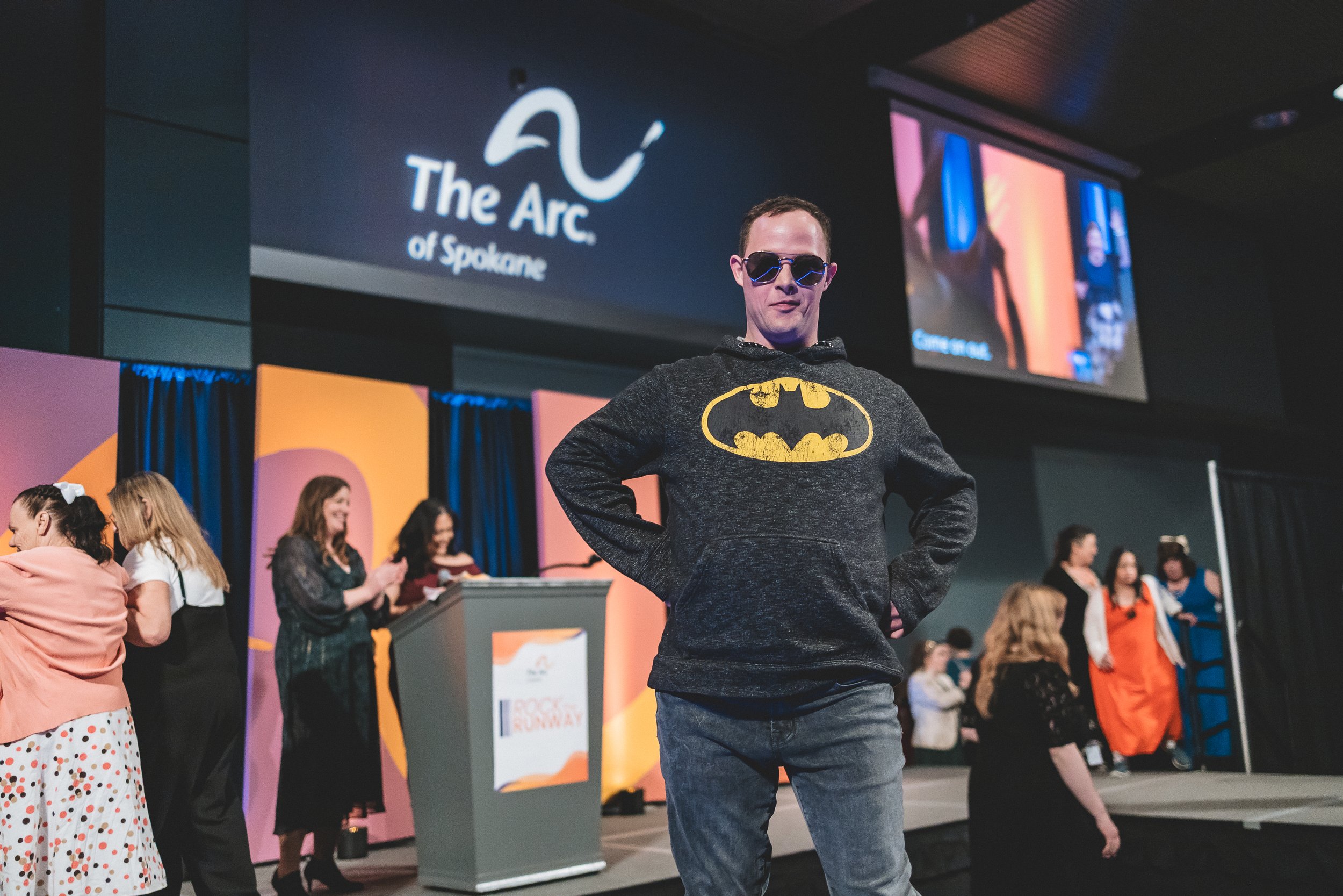 An image of a Rock the Runway model wearing sunglasses and a batman sweatshirt. They pose on the stage in front of The Arc of Spokane's logo.