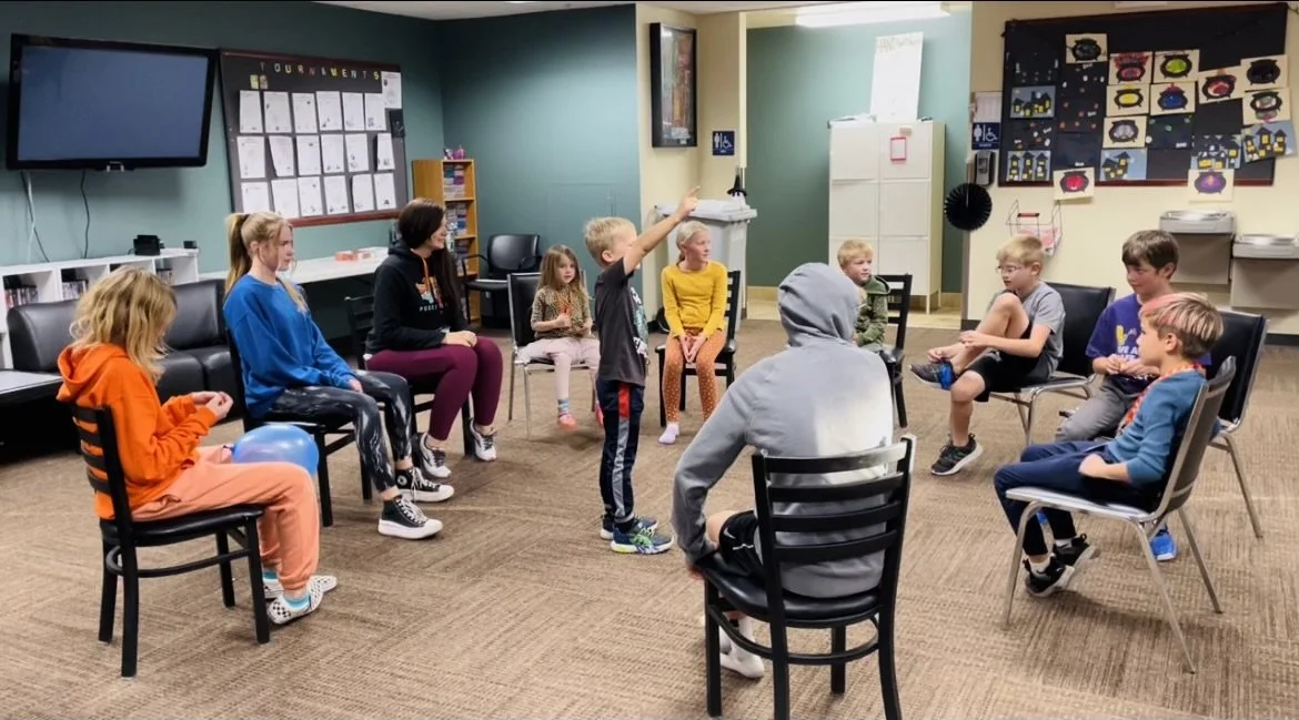 A group of children and teens sit in a circle on chairs as one child stands in the center to lead a game or discussion.