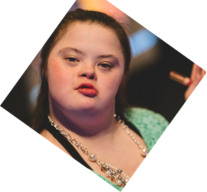 A glamour close up of a femme presenting Rock the Runway model. They give a dramtic smouldering look while wearing a green floral top with an assortment of beaded necklaces