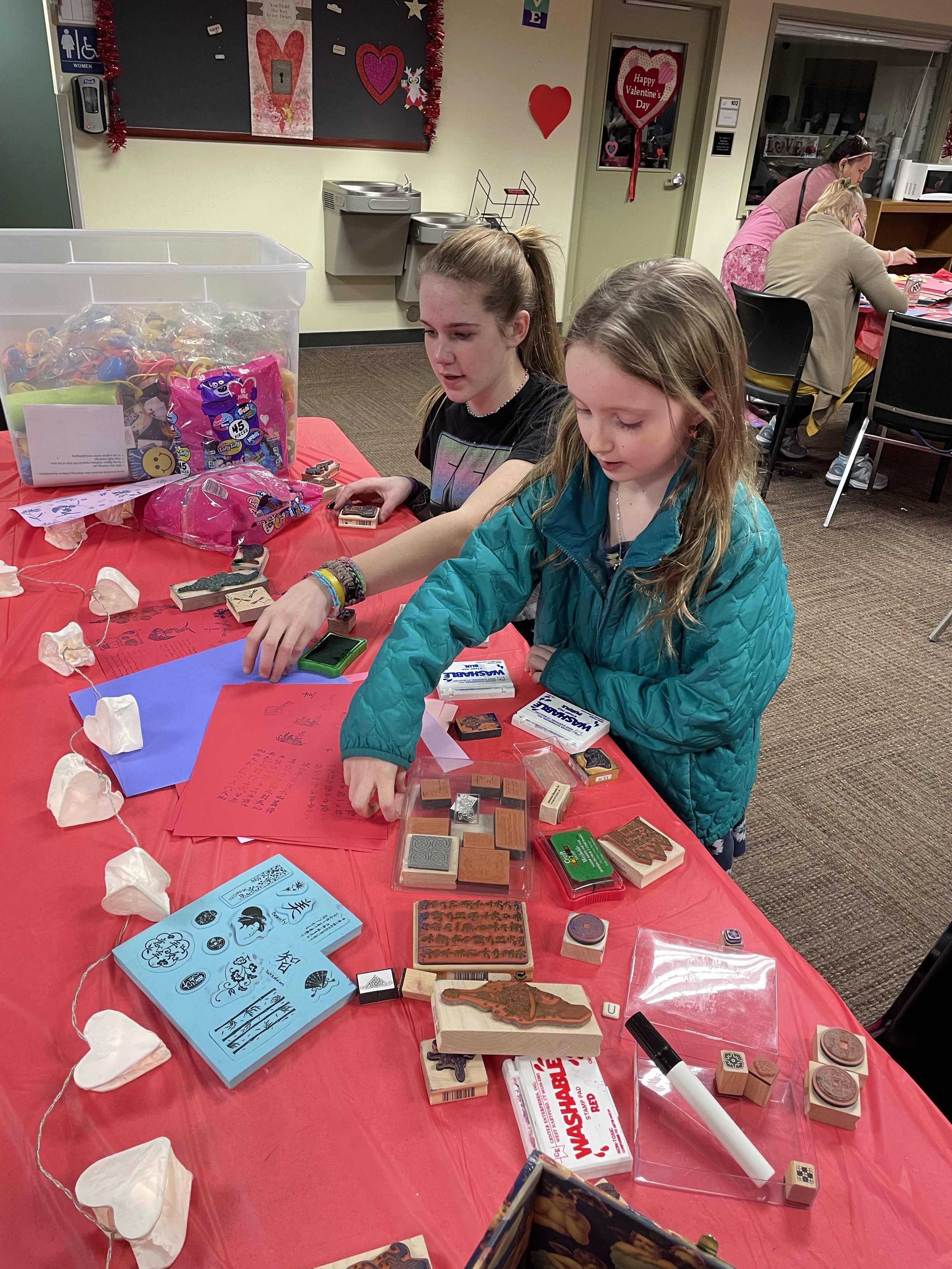 Two girls stand at a table covered in a red cloth, carefully selecting and using rubber stamps on colorful paper at a workshop.