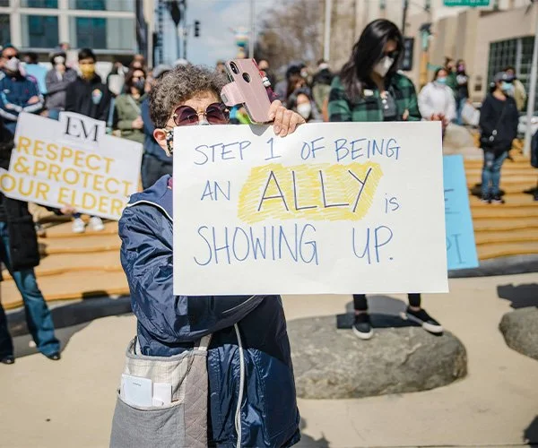 Image of a women holding up a sign at a protest that says "Step 1 of being an ally is showing up."