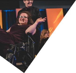 Image of a femme presenting model being pushed in their wheelchair. With a hige smile on their face, they give the audience a delicate princess wave. They are wearing a burgendy velvet dress.