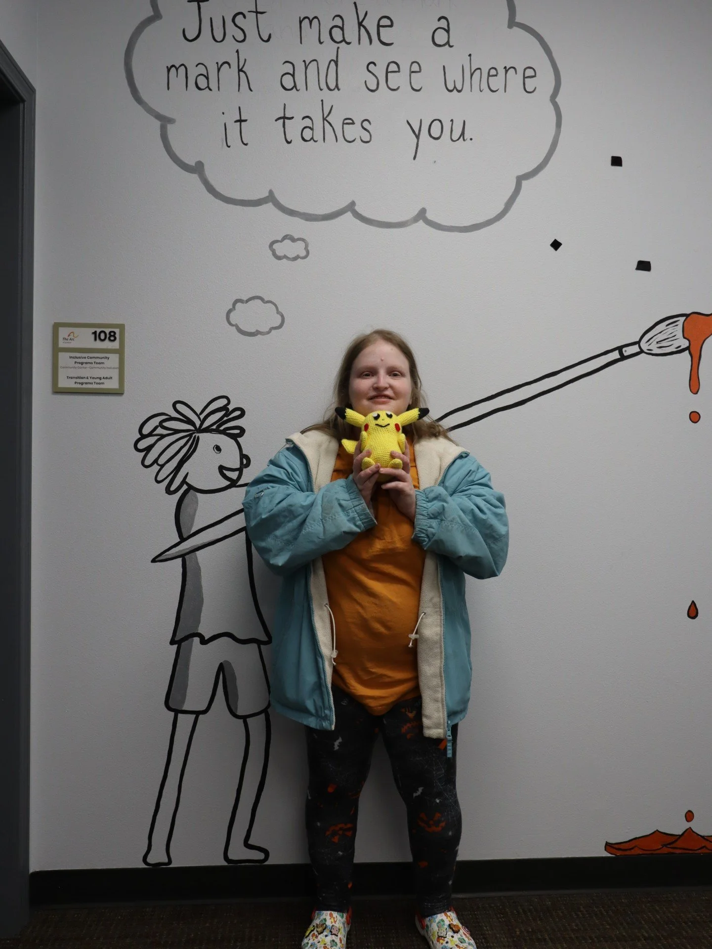 Cubley, a member of The Arc of Spokane community, smiling while holding her yellow hand-knitted Pikachu. She stands in front of a wall mural that reads, "Just make a mark and see where it takes you.