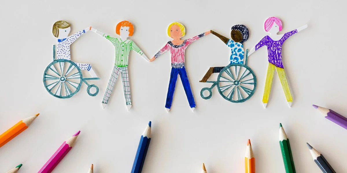 Five hand‑drawn paper figures, including two using wheelchairs, hold hands above a semicircle of colored pencils on a light background.
