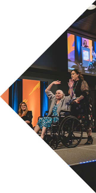 An image of a femme presenting model wearing a blue dress and gray cardigan. As they are pushed across the stage in their wheelchair, their hands are held high in celebration
