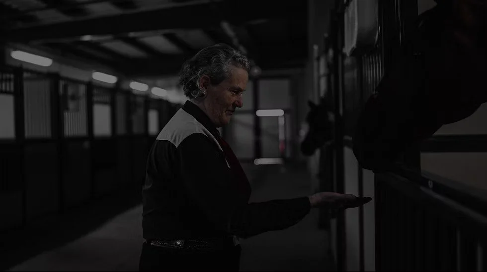 Temple Grandin stands in a stable and holds her hand out to a horse in a stall. There are several other horses in the background. The image has a dark gray overlay and was provided by https://www.templegrandindocumentary.com/