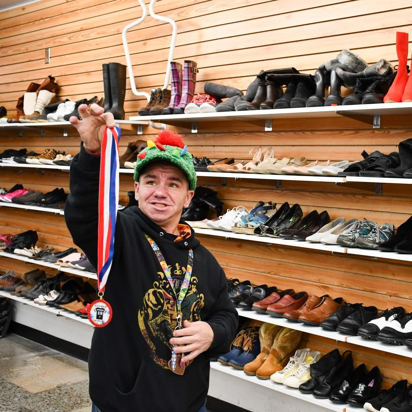 A member of The Arc of Spokane's Community Center stands in front of a shoe rack in The Arc Thrift Store's Spokane Valley Location. He smiles wide while wearing a fuzzy green hat with colorful felt spikes. He holds up a medallion he thrifted.