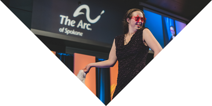 A femme presenting model from Rock the Runway poses elegantly with a huge smile on their face. The model is wearing a sparkling dress in a deep purple shade with a pair of bright pink sunglasses