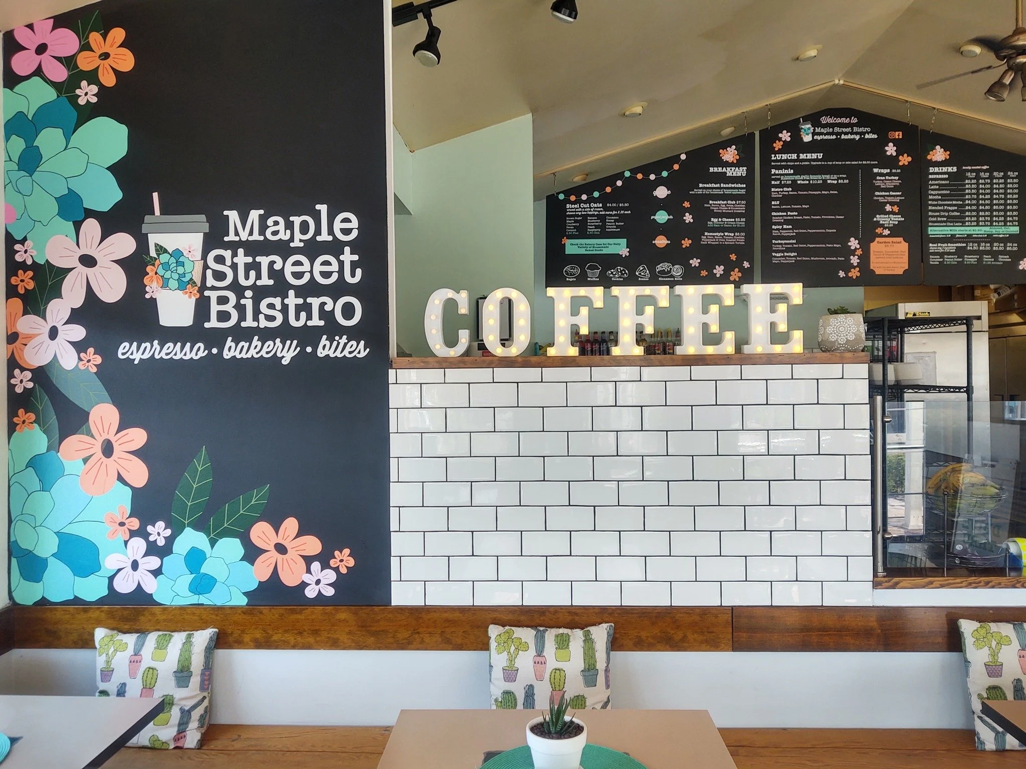 Interior of Maple Street Bistro with floral wall art, menu boards, and a lit COFFEE sign above white tile.