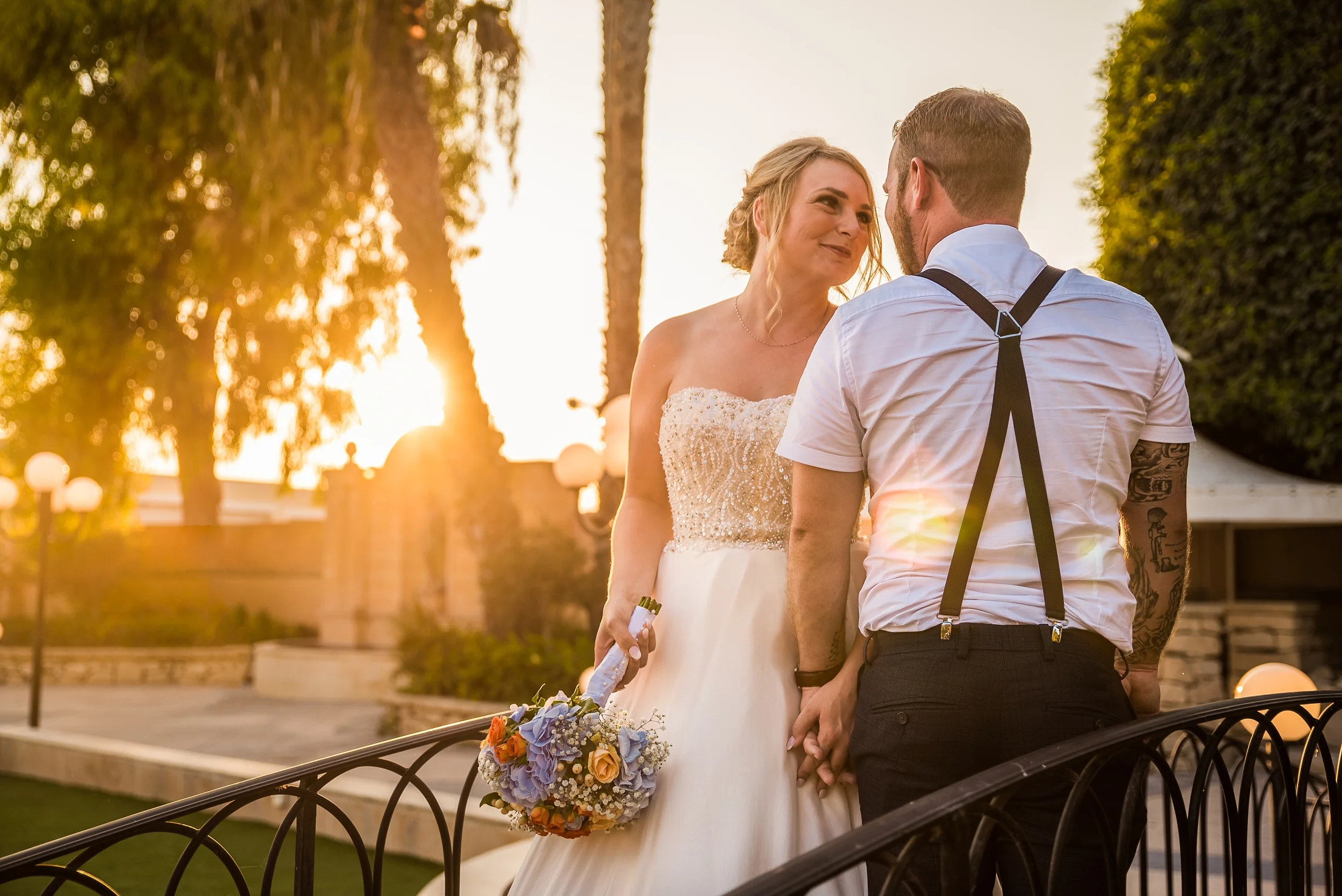 A bride and groom holding hands and gazing at each other during sunset outdoors, with trees and decorative lamp posts in the background.