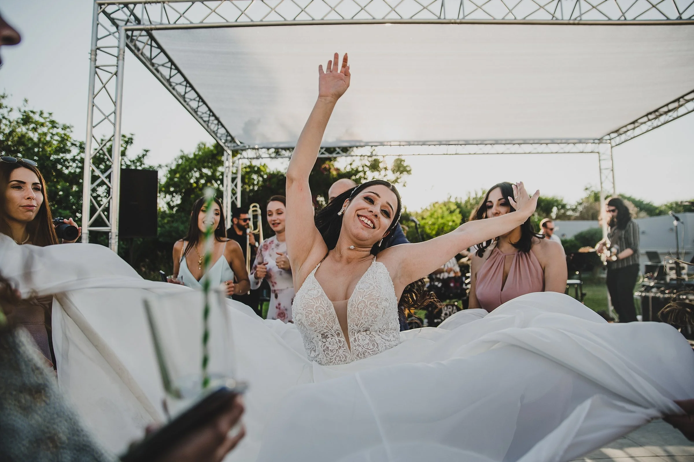 A bride in a white wedding dress dancing joyfully at her outdoor wedding reception with guests in the background.