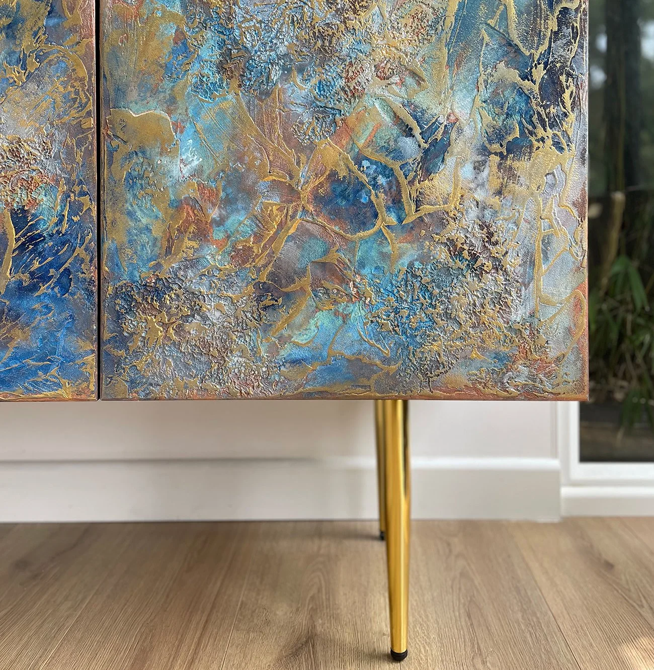 Close-up of a Midnight Galaxy sideboard door featuring heavy cerulean and copper textures, transitioning into a sleek, straight gold-finish furniture leg on light oak flooring.
