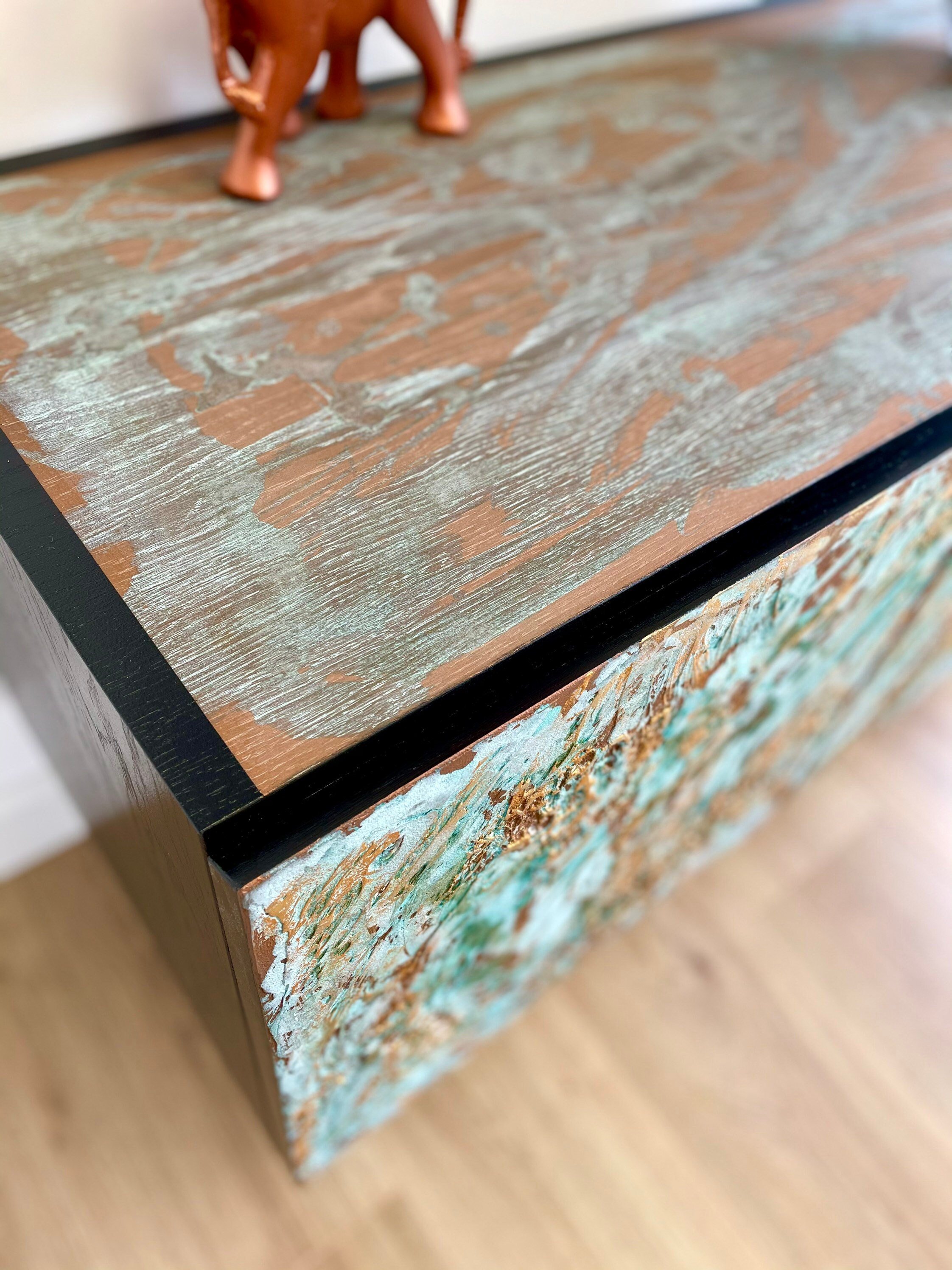 A close-up of a textured wooden table, with a weathered, rustic finish in shades of brown and teal, highlights its bespoke charm.