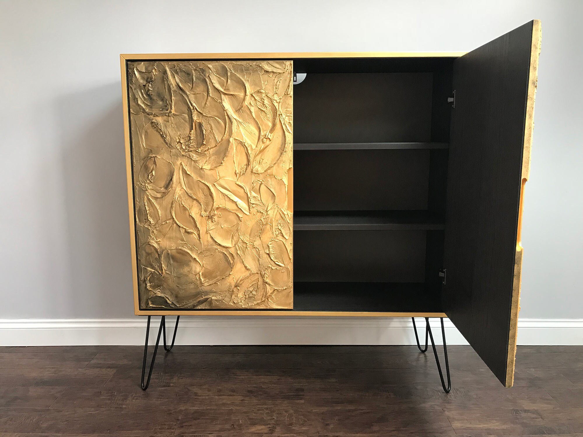 A bespoke wooden cabinet features a textured gold-leaf front on the left, while an open black door on the right reveals shelves inside. It stands on sleek metal hairpin legs.