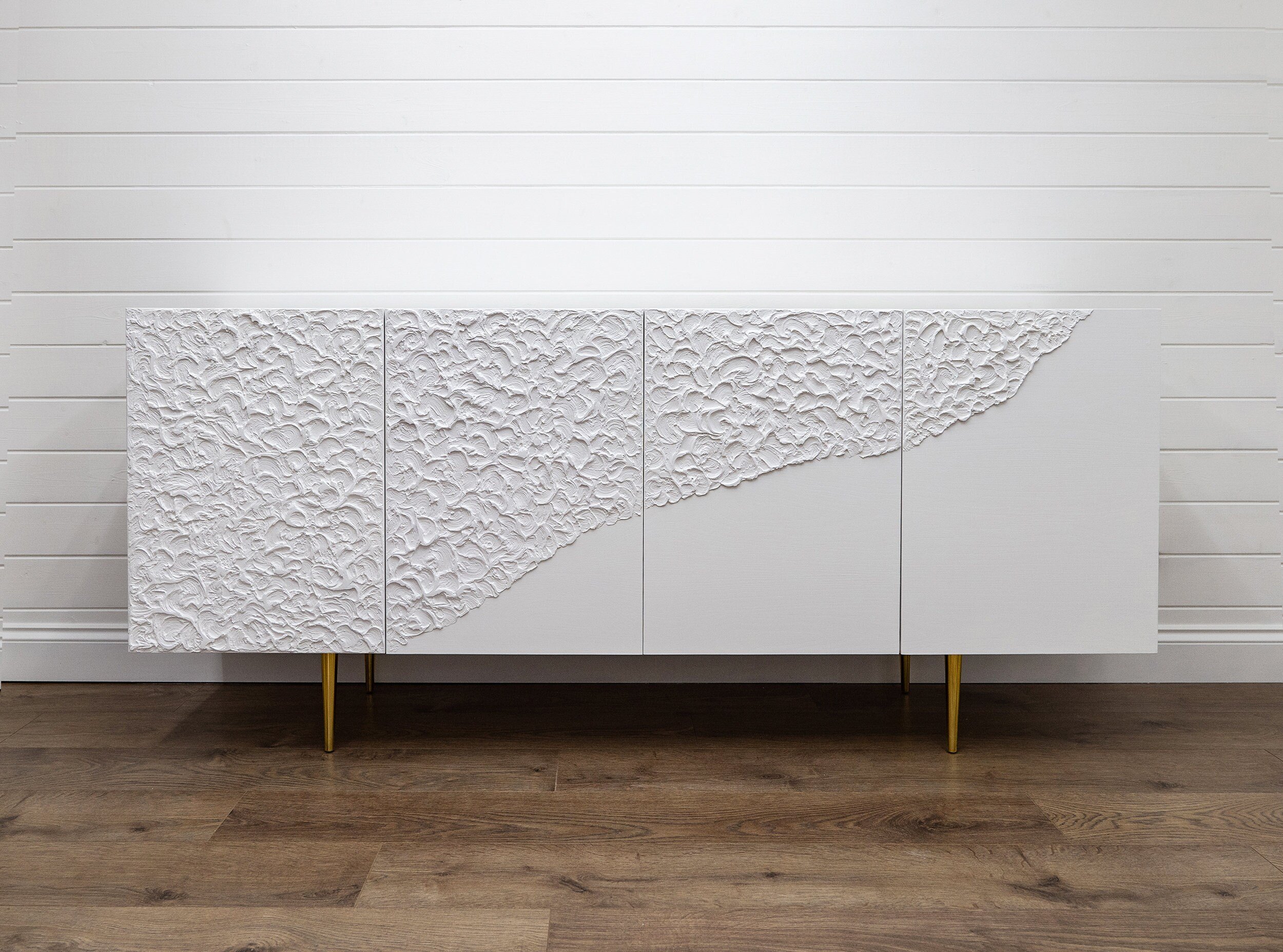 A modern white custom-made sideboard features textured, wave-like patterns on the front. This bespoke furniture piece stands on sleek, gold legs.