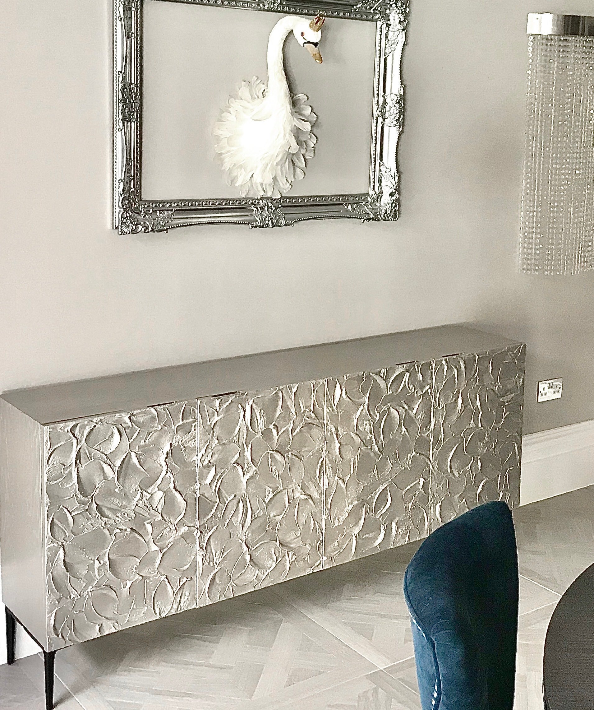 A room featuring a bespoke silver floral-patterned sideboard, a teal velvet chair, and a swan sculpture mounted in a decorative frame on the wall above.