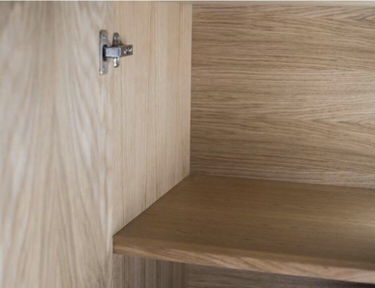 An empty wooden cabinet with light brown wood grain, featuring a metal hinge on the left side and a single shelf inside. The cabinet interior and the shelf have a natural finish.