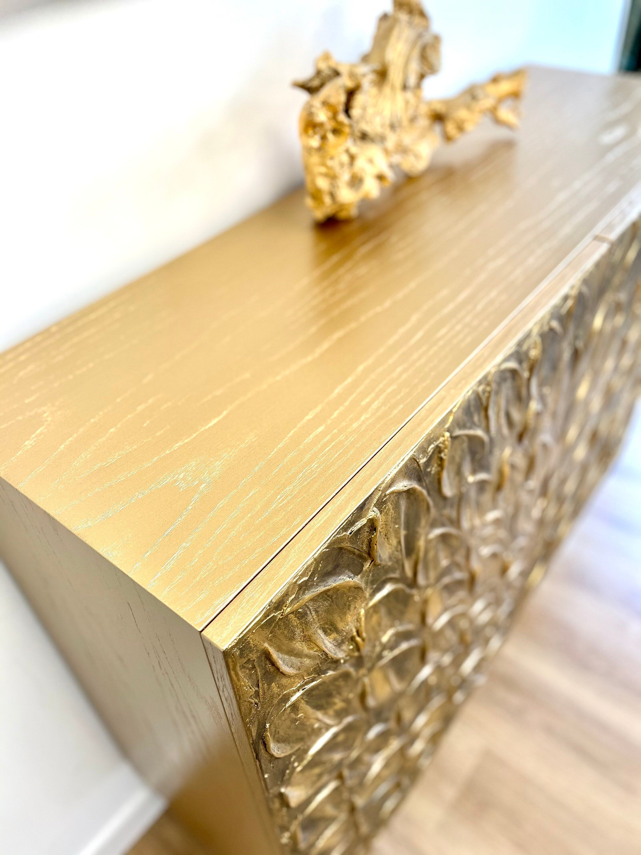 Golden bespoke console table with a textured floral design on the front and a shiny golden surface. A small, abstract gold sculpture is placed on top.