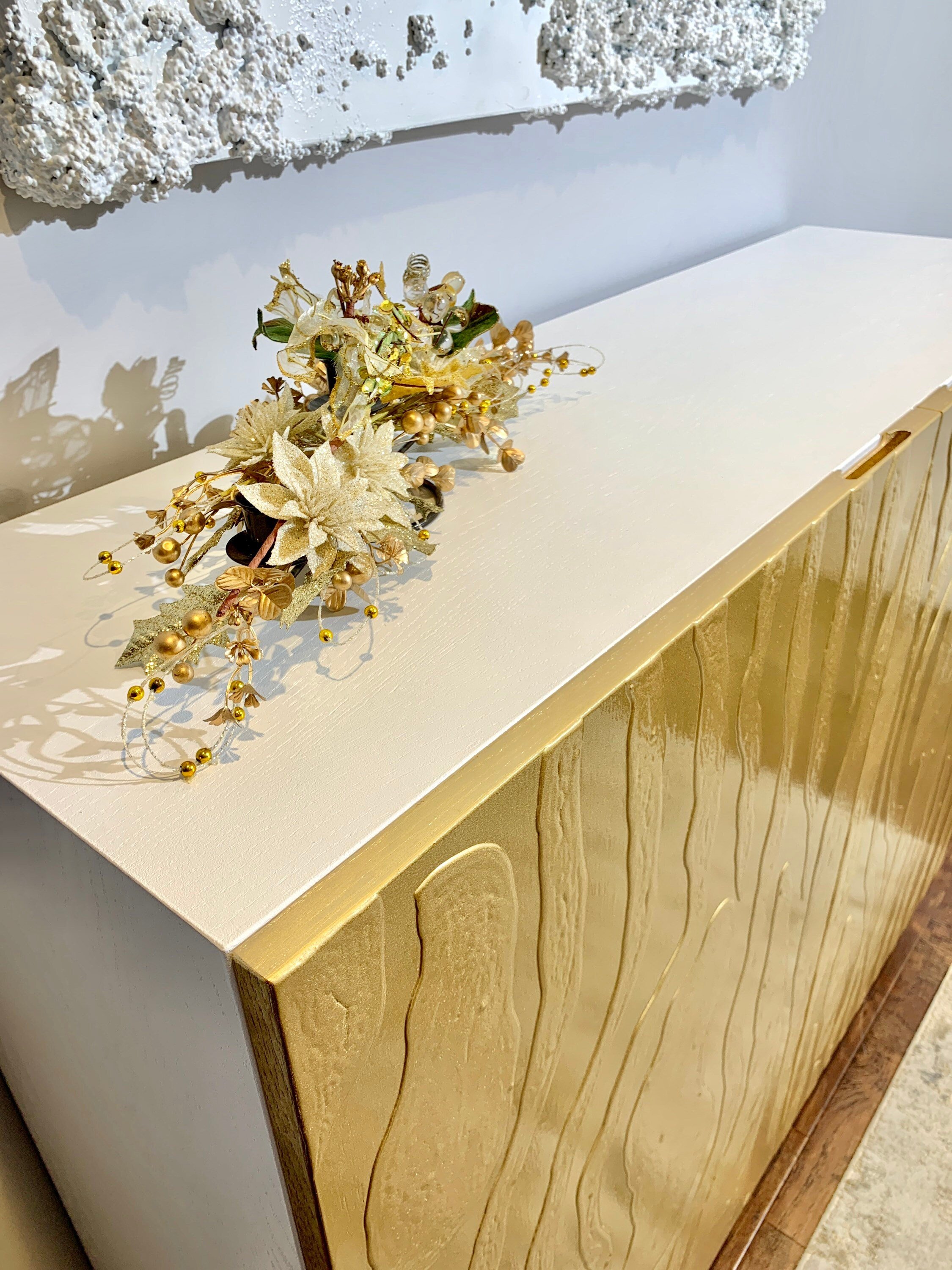 A modern cabinet boasts gold textured doors and a smooth white top, adorned with decorative gold and white flowers and beads.