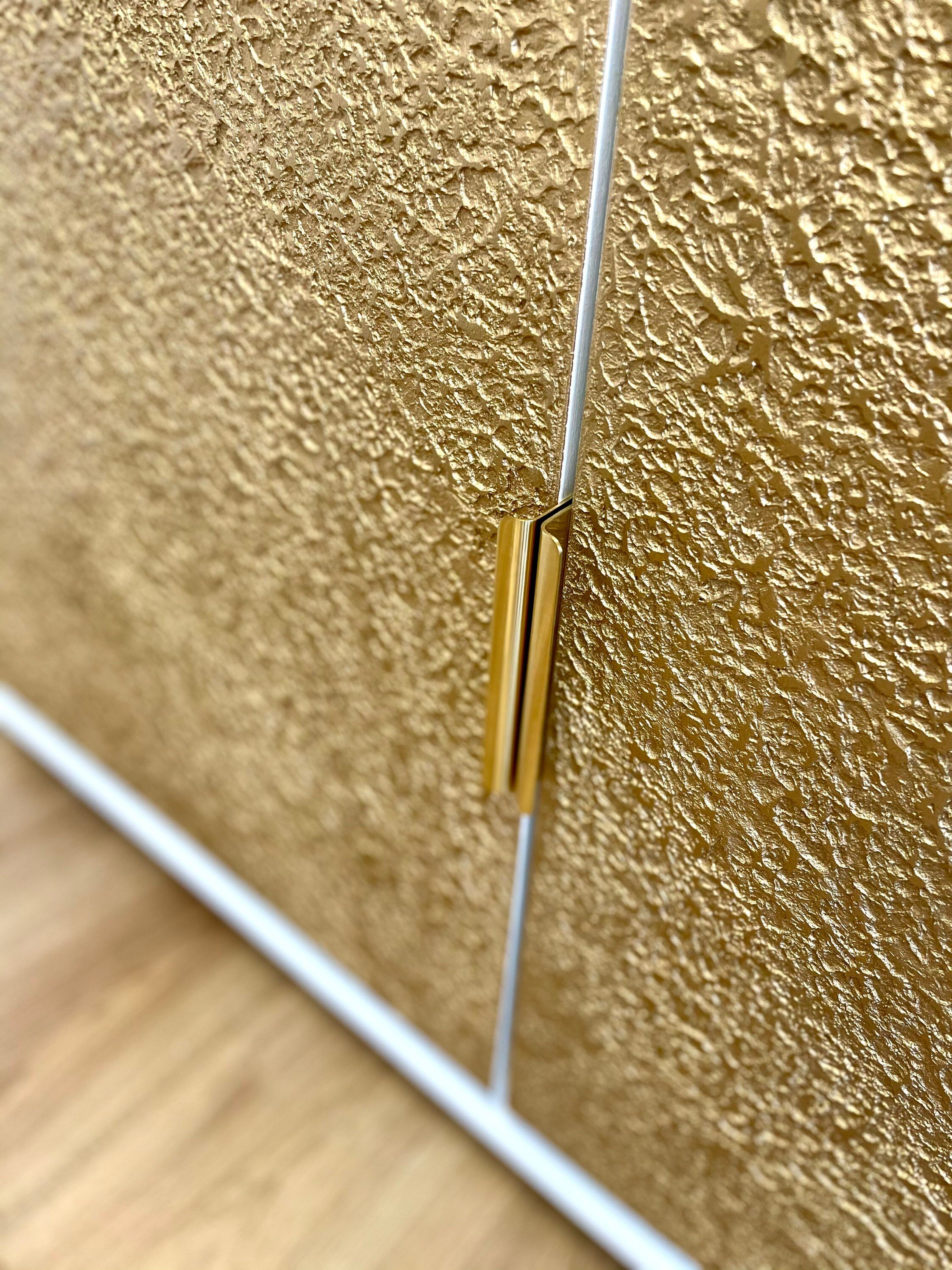 Close-up view of a textured, golden cabinet door on a wooden floor, featuring a sleek, vertical metallic handle in the center.