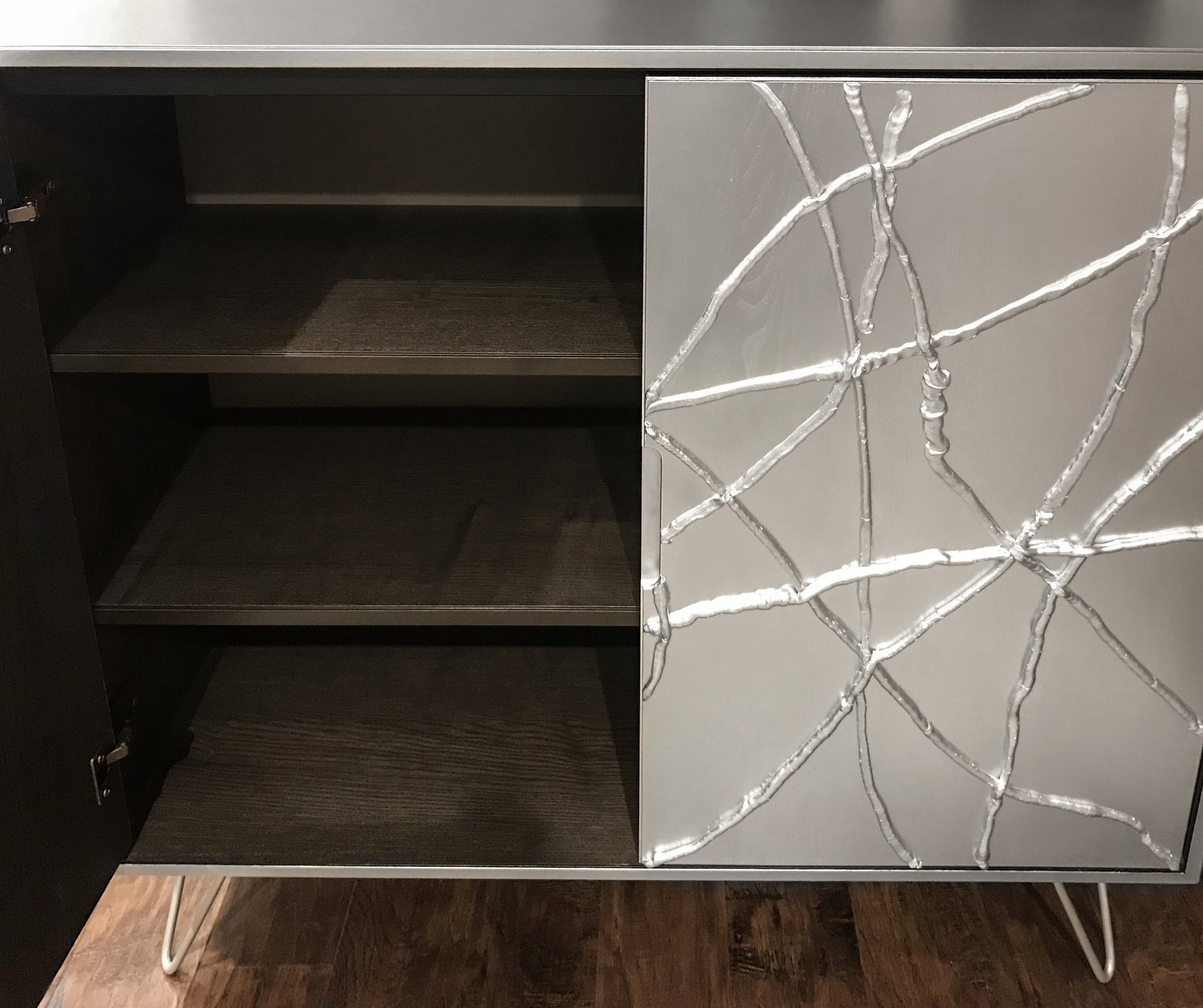 A modern metallic, custom-made sideboard with an open door reveals two empty, dark wooden shelves. The right door features an abstract silver wire design that adds texture.