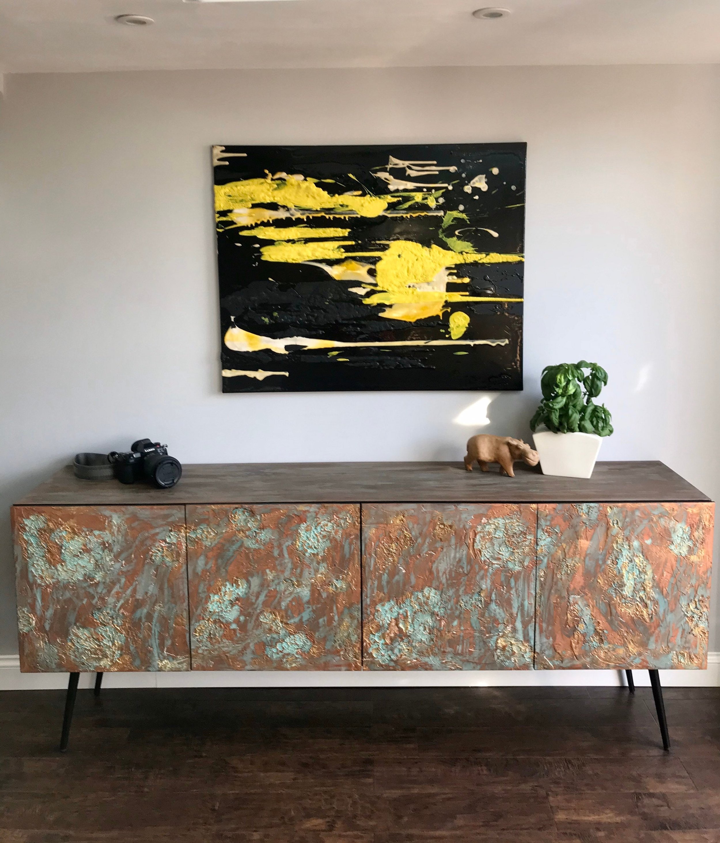 A custom-made sideboard with a distressed wood finish stands against a light gray wall. A large abstract painting with black and yellow drips hangs above it.