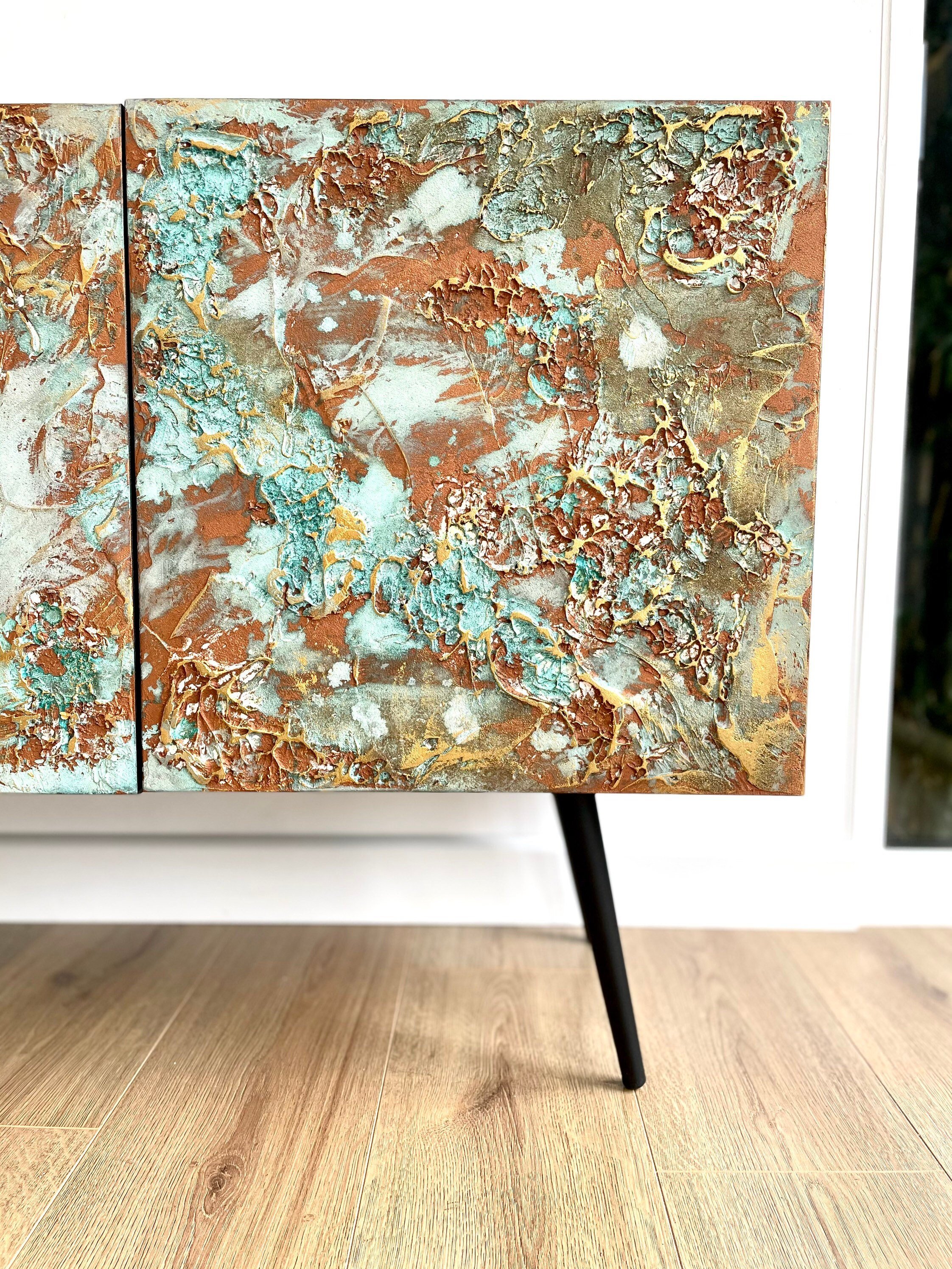 A close-up of a bespoke cabinet with two doors featuring textured art in earthy tones of brown, teal, and gold. The cabinet stands on sleek black legs.