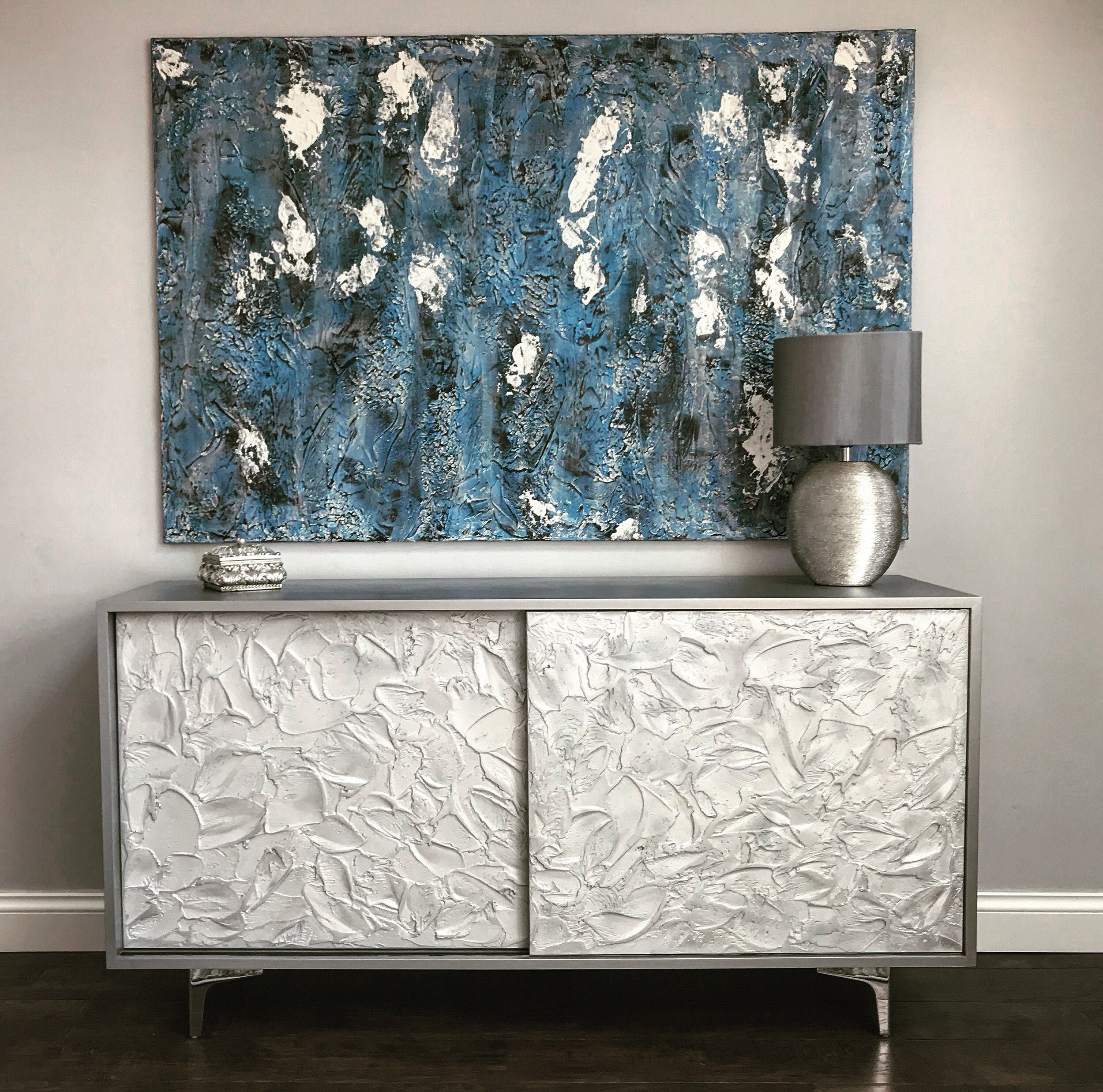 A custom-made sideboard with textured, white sliding doors stands against a gray wall. A blue abstract painting hangs above it.