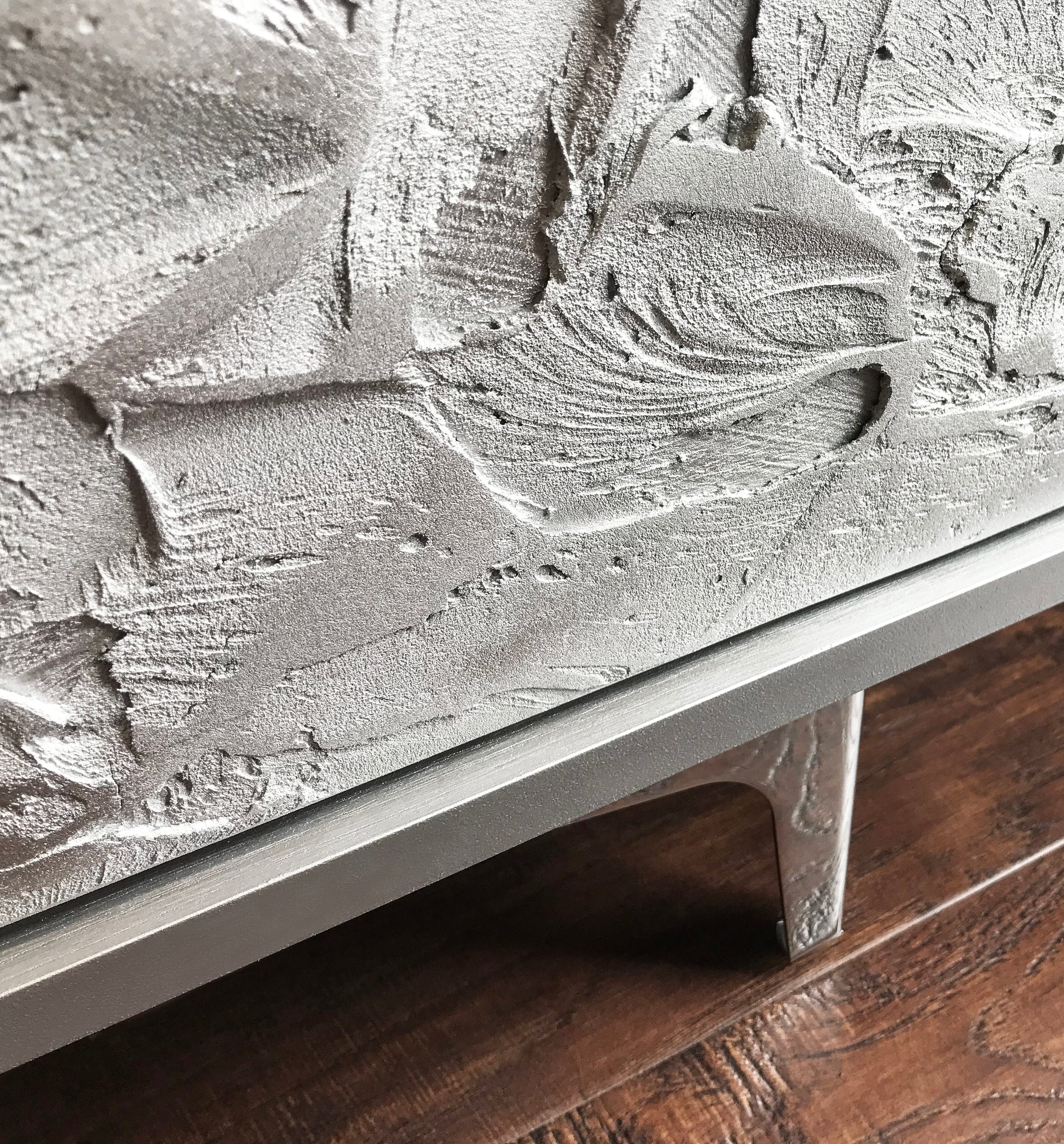 A close-up of a textured silver-gray surface with abstract patterns, mounted on a sleek, bespoke furniture stand. The custom-made sideboard resembles a mix of smooth and rough strokes.
