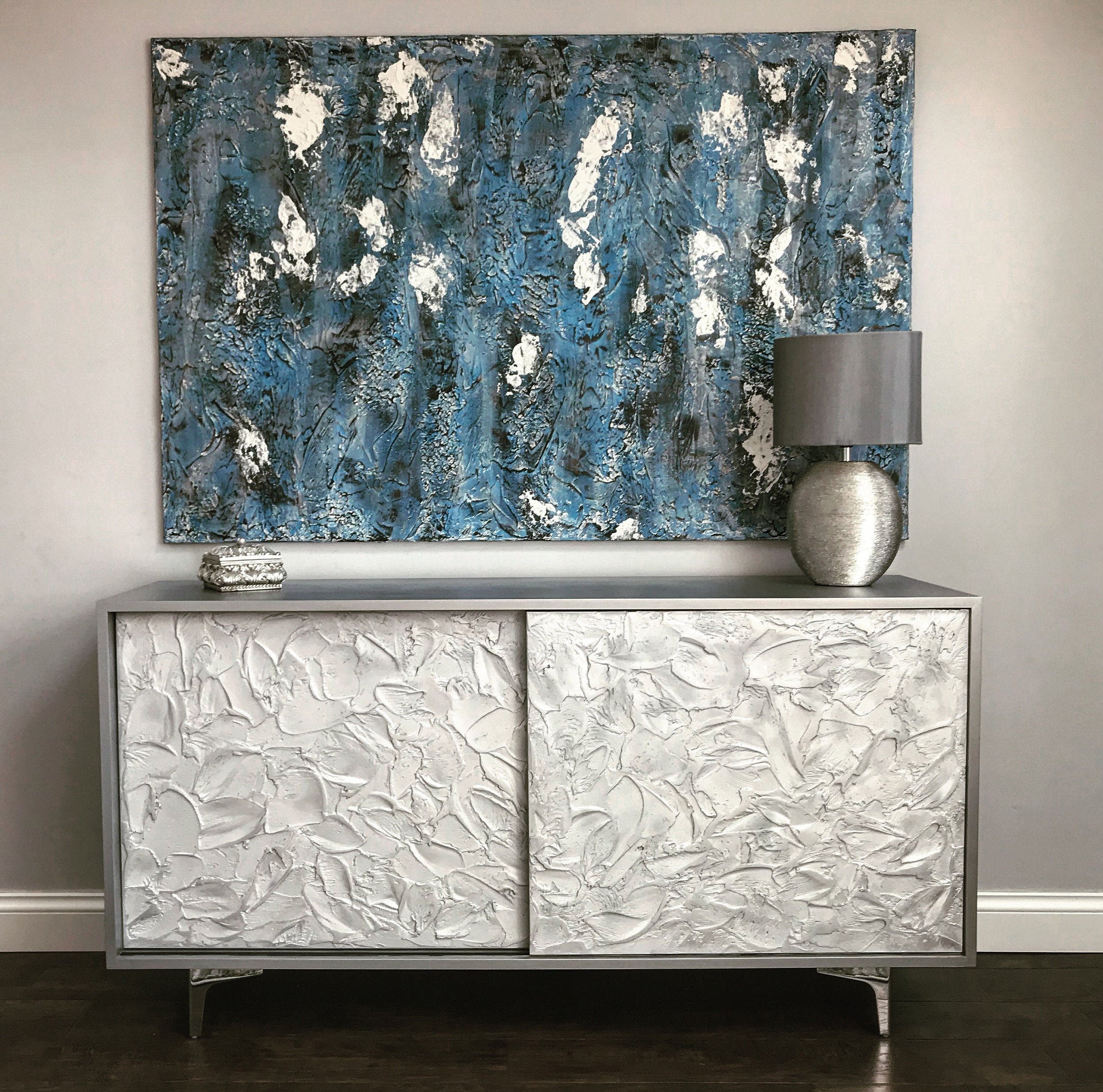 A custom-made sideboard with textured white surfaces and abstract designs stands in front of a wall. Above it hangs a large, blue and white abstract painting.