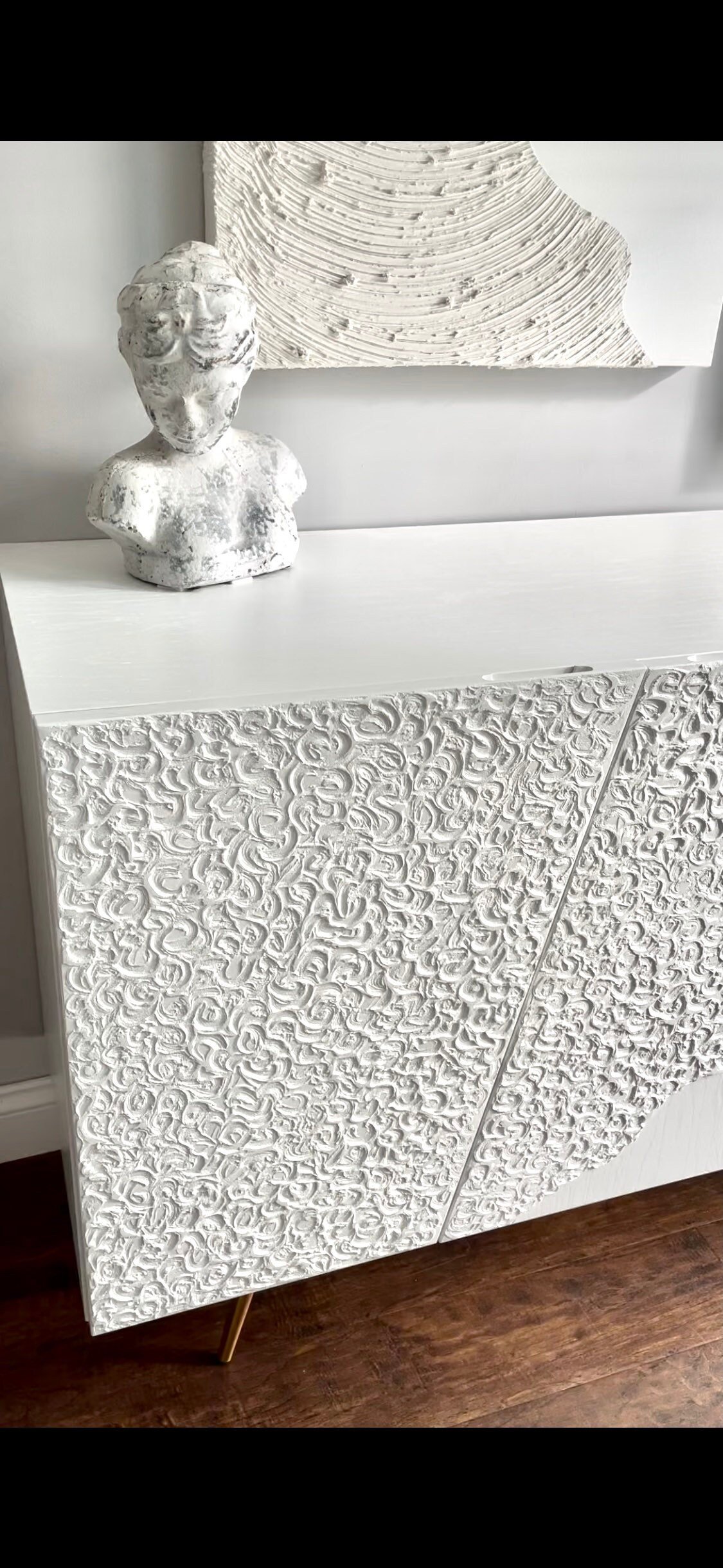 A custom-made sideboard with a decorative pattern stands on a wooden floor. On top, theres a white bust sculpture. A piece of art with a similar texture is partially visible above.