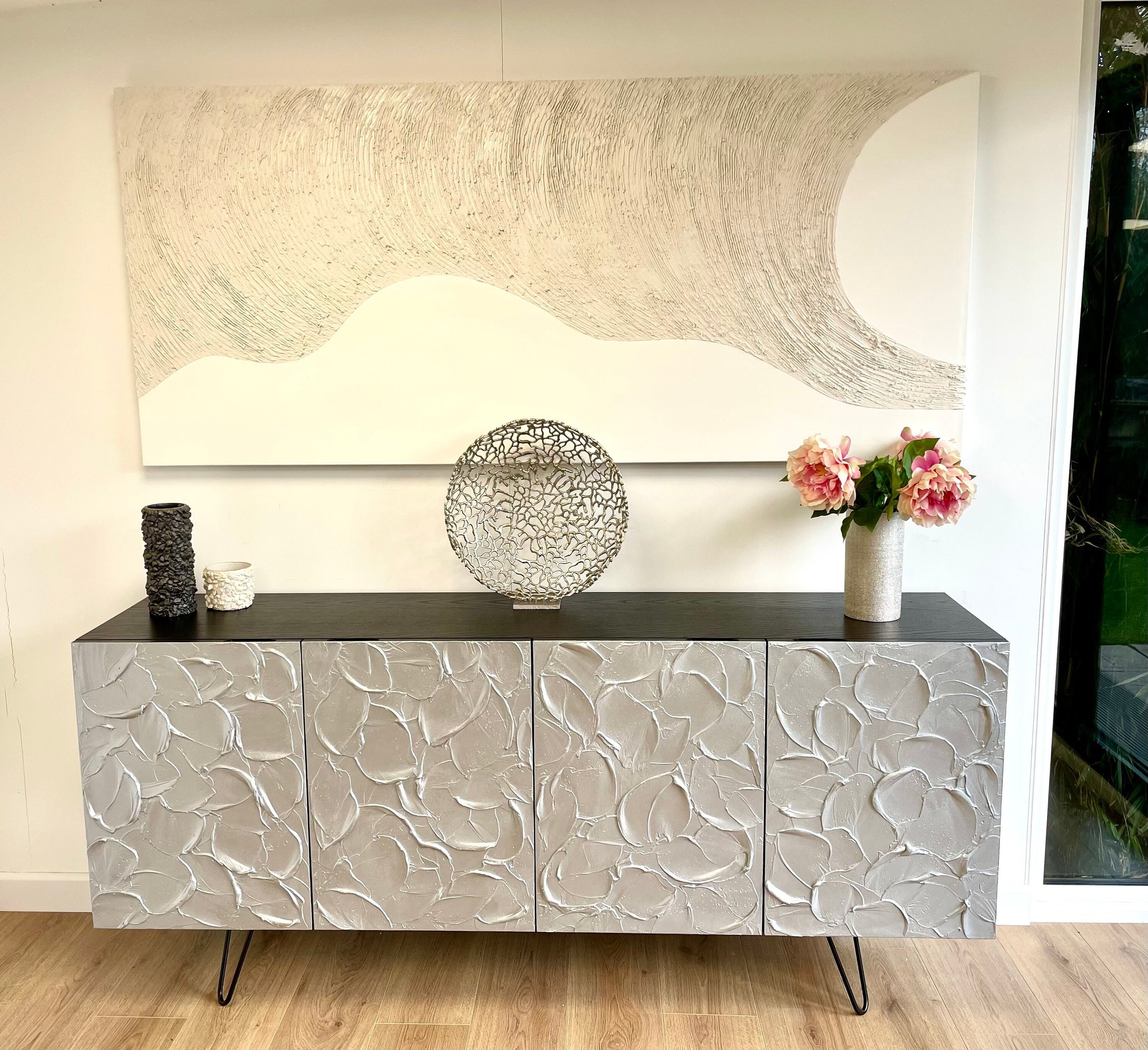 A modern room showcases bespoke furniture with a textured sideboard adorned in floral patterns. A large abstract painting in neutral tones hangs above.